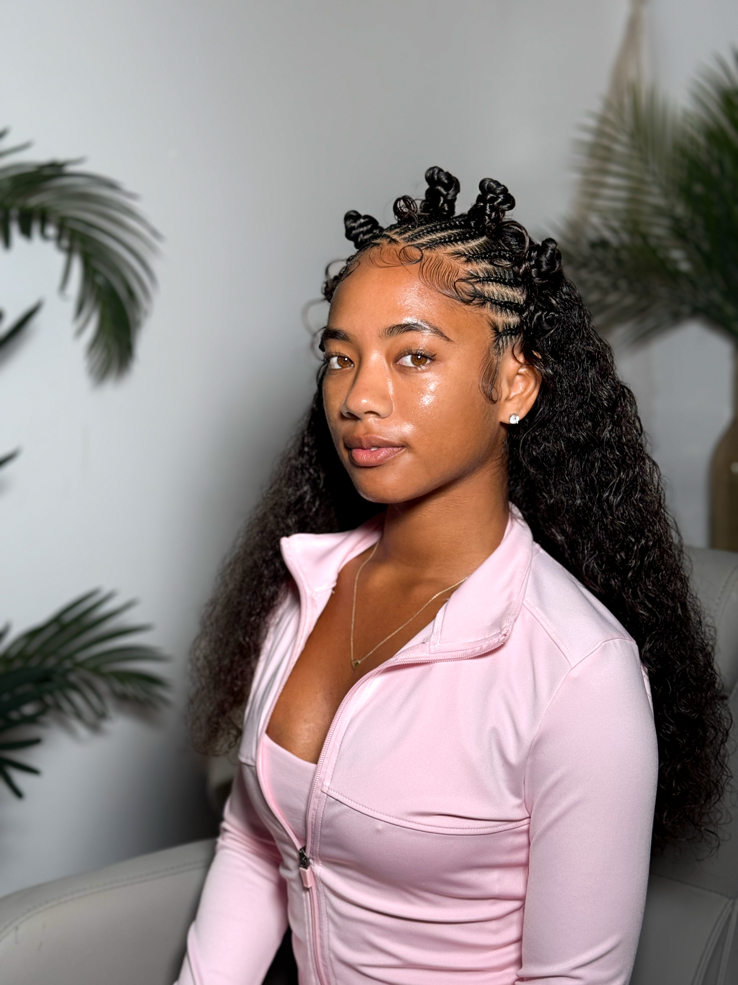 A young woman with medium brown skin, curly long hair styled in small buns with cornrows, wearing a light pink zip-up top, seated near a white wall with green foliage on the sides.