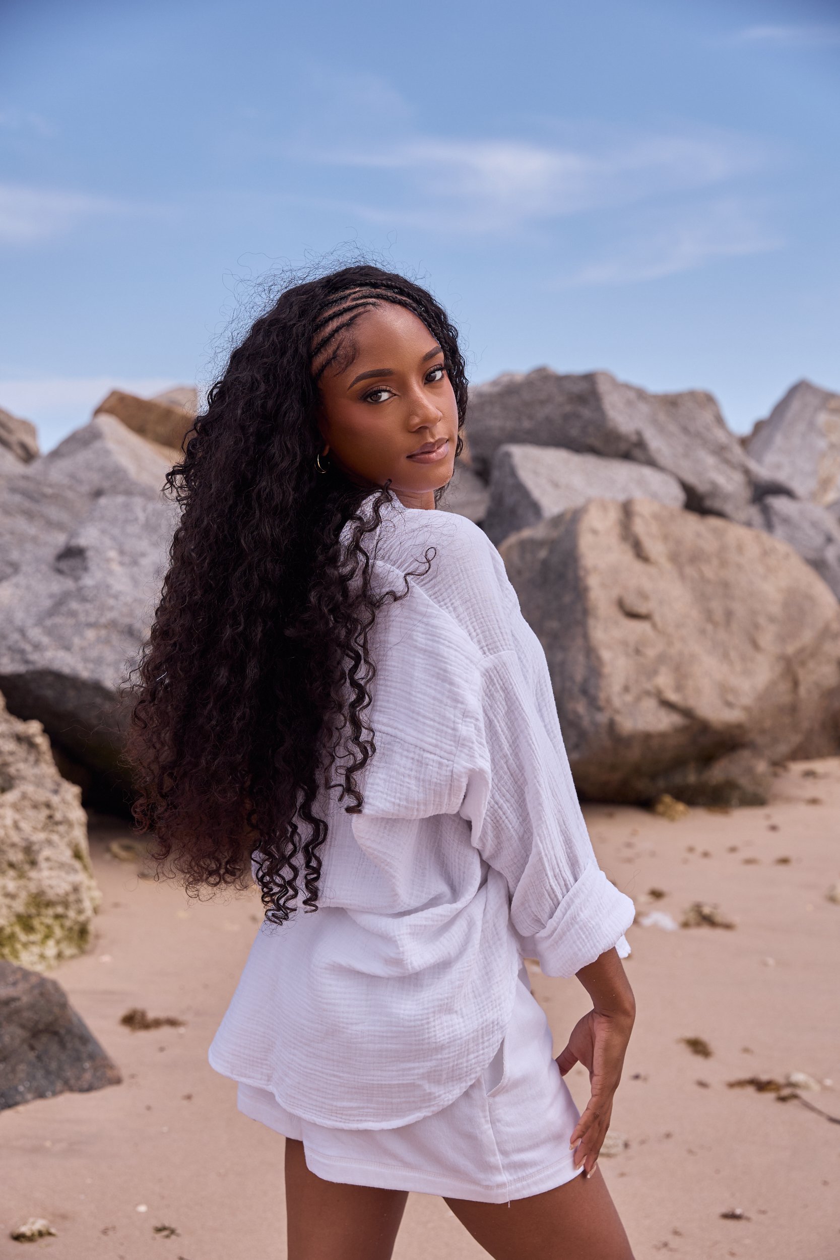 A woman with long curly hair and a white outfit standing on a rocky beach with clouds in the sky.