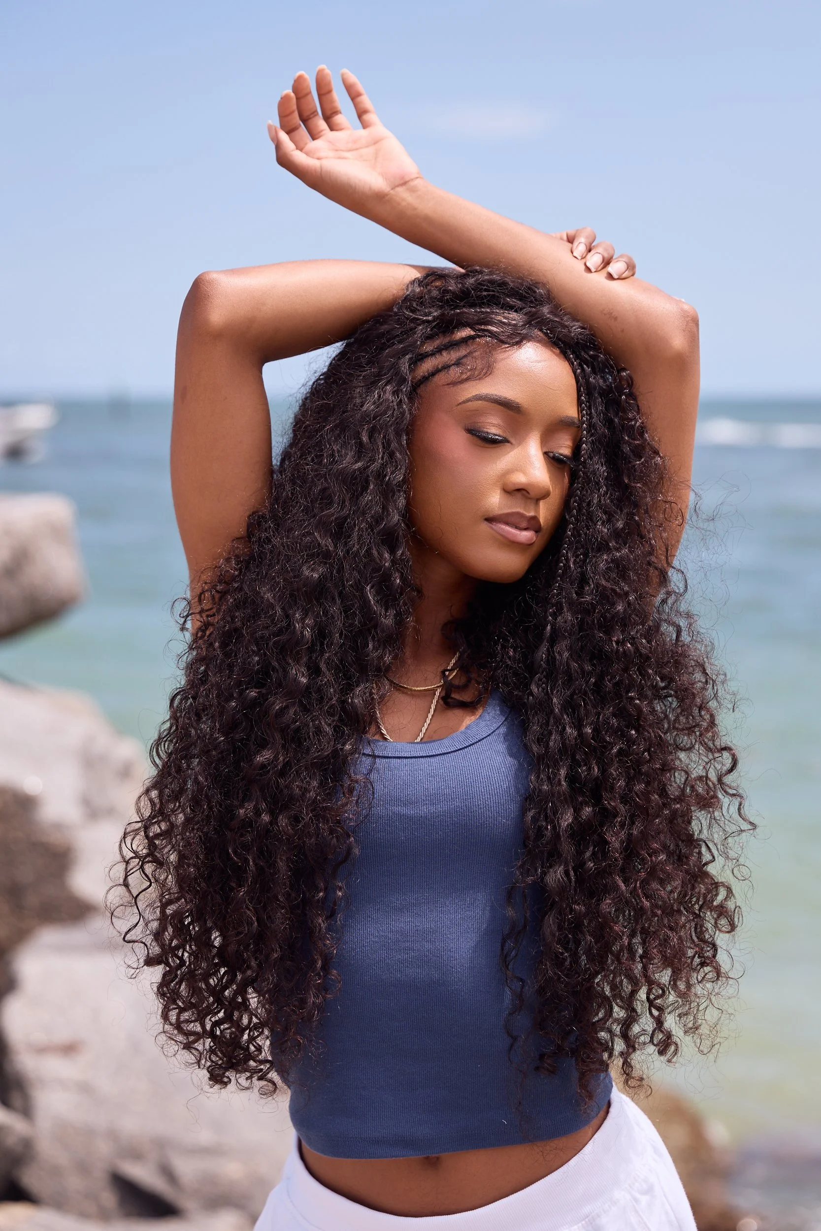 A young woman with long, curly hair standing on a beach with rocks and the ocean in the background, wearing a blue sleeveless top and white shorts, with her arms raised and hands touching her head, enjoying a sunny day.
