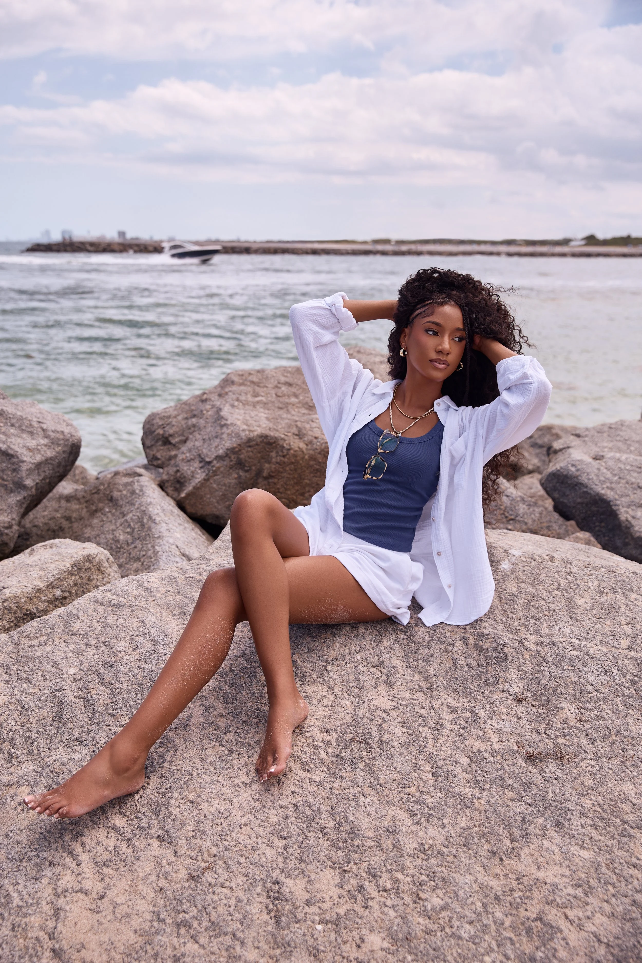 A woman with curly hair sitting on rocks at the beach, wearing a blue top, white shorts, a white shirt, and jewelry, with water and sky in the background.