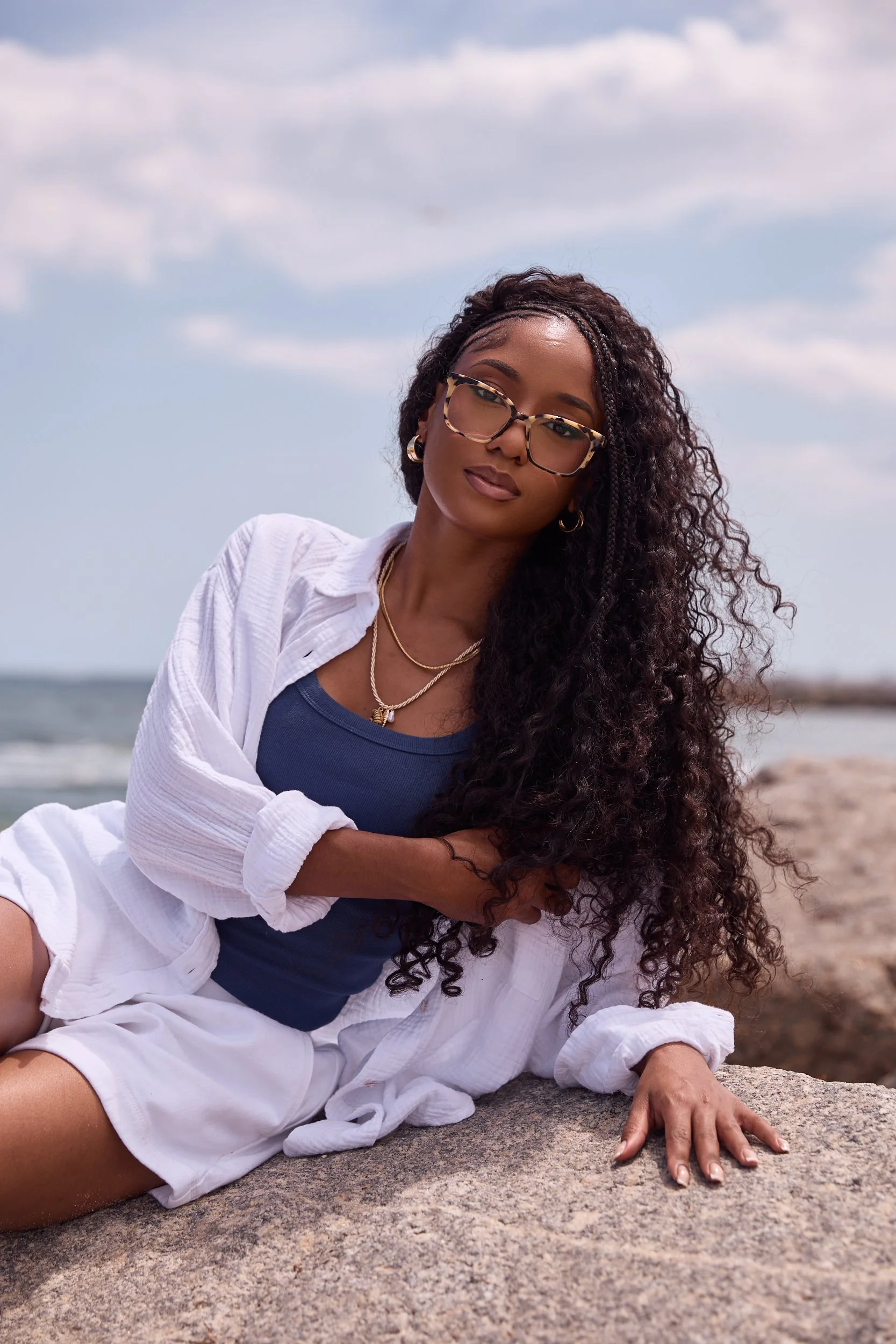 A woman with long curly hair and glasses sitting on a rock at the beach, wearing a navy blue top, white shorts, and a white shirt, with her hand resting on the rock and her face turned slightly away from the camera.