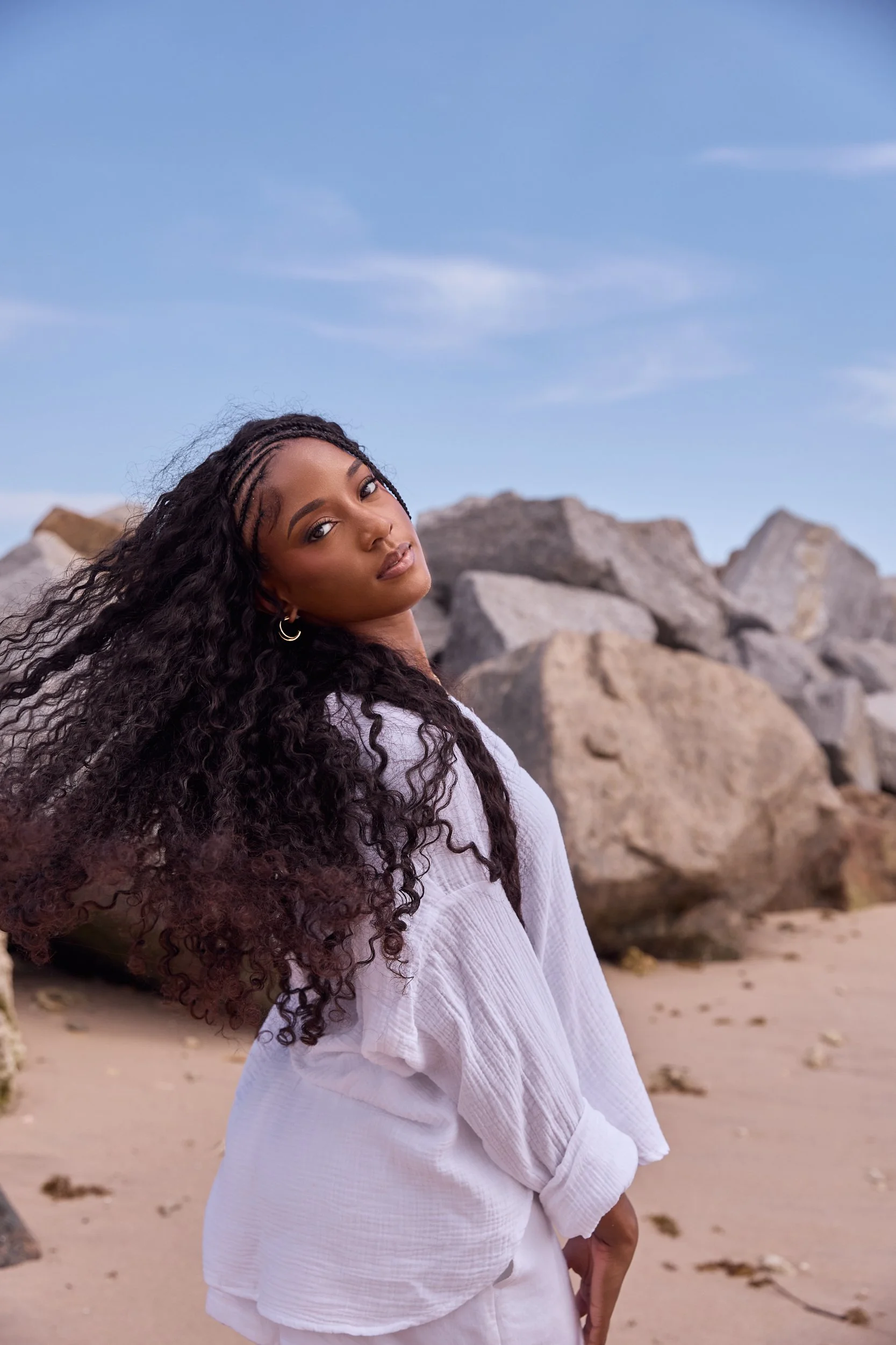 A woman with long, curly hair standing on a beach with rocks in the background, wearing a white top and white pants, under a blue sky with a few clouds.