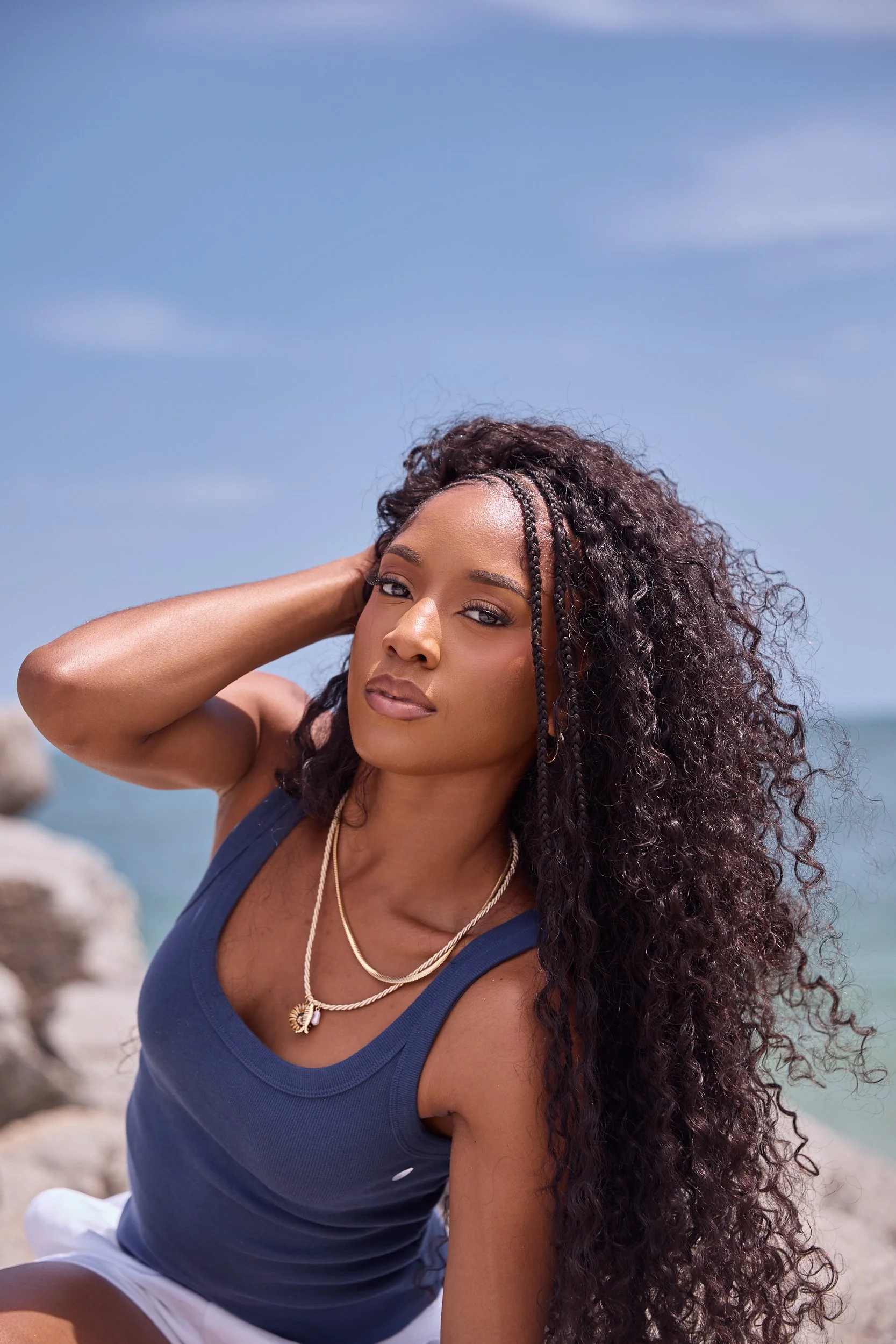 Close-up of a young woman with long, curly hair wearing a navy blue tank top and white shorts, sitting outdoors near the water on rocks, with a blue sky in the background.