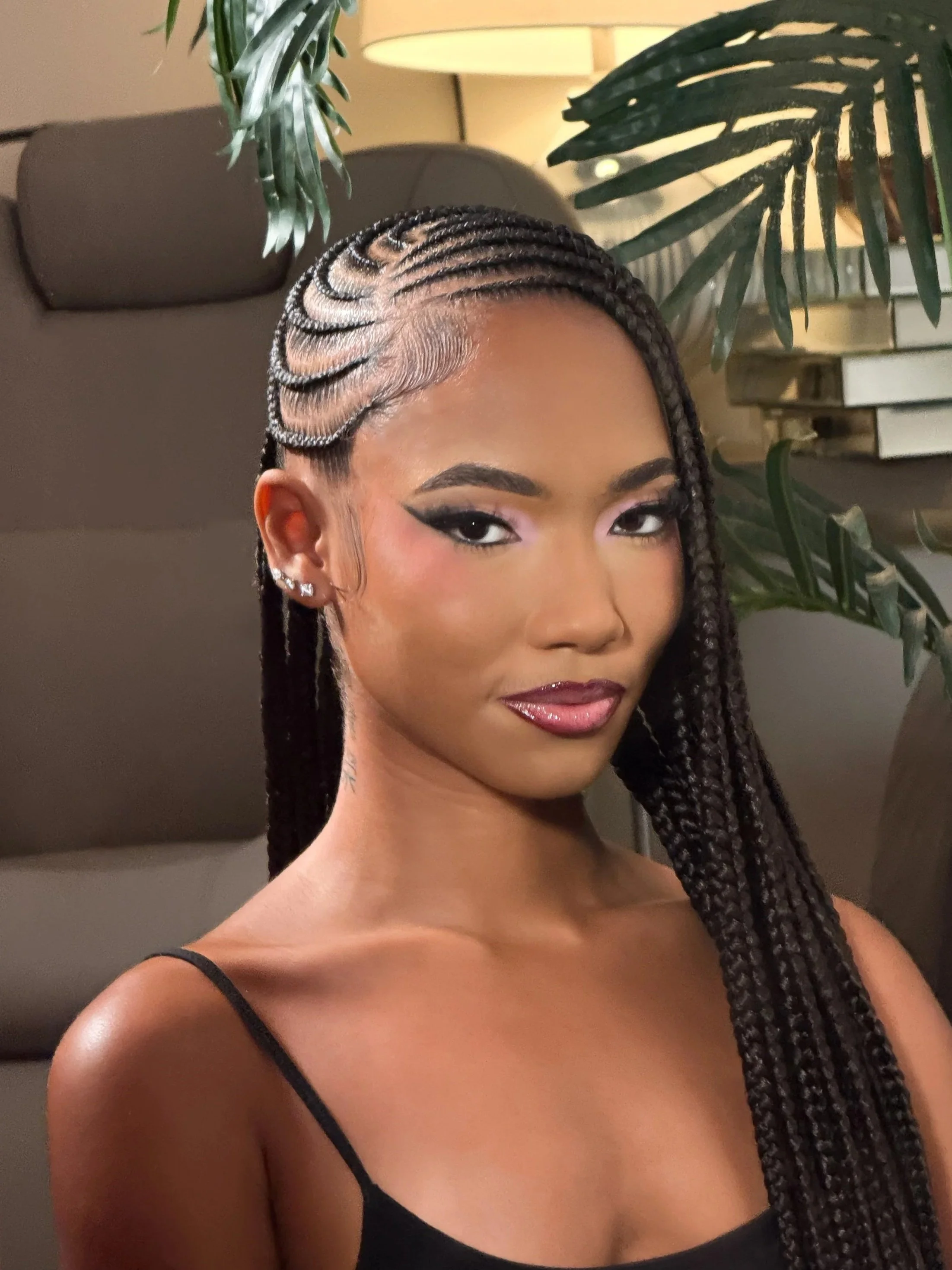 Close-up of a woman with braided hair, wearing black straps, makeup, earrings, and with a background featuring green plants and a beige lamp.