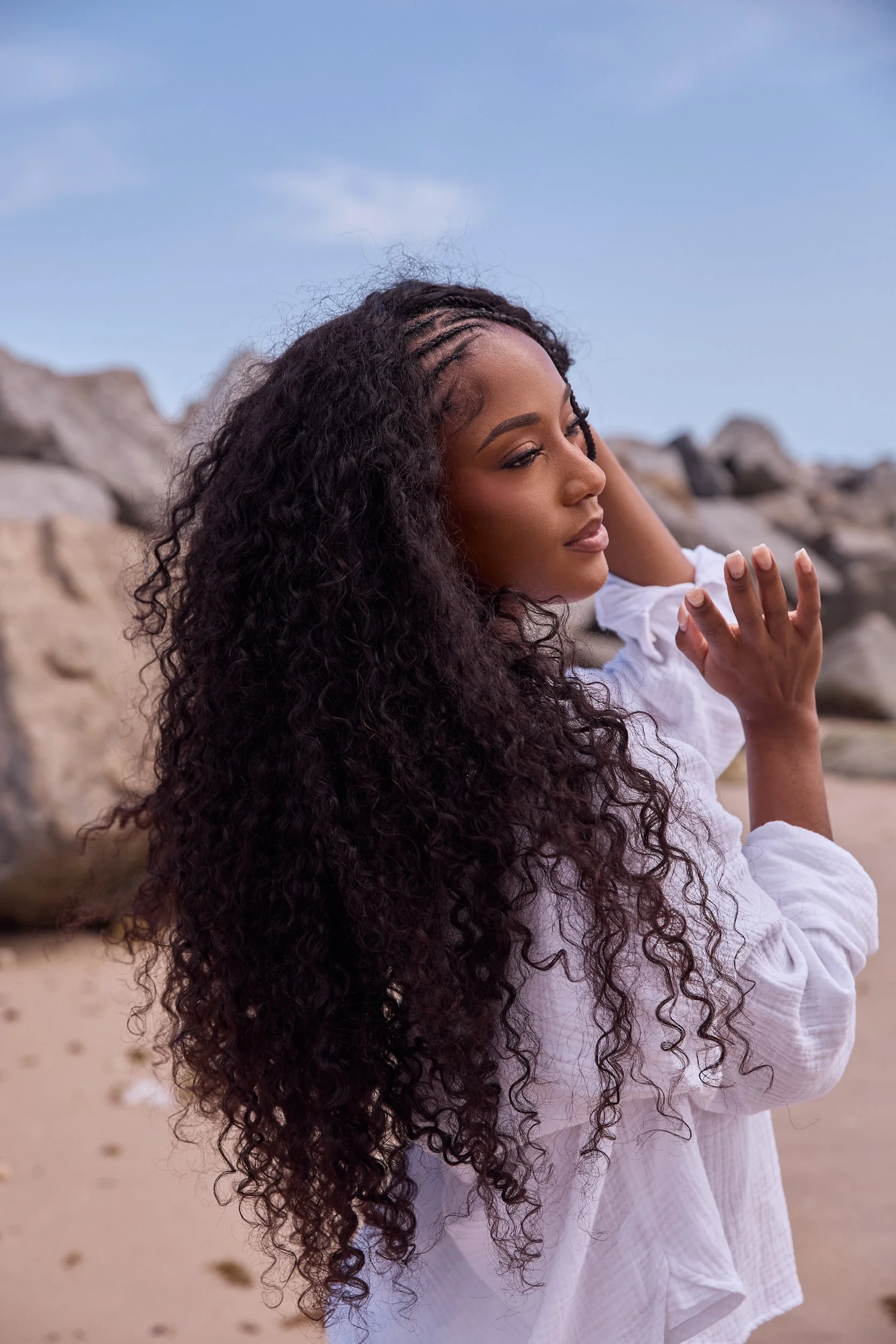 A woman with long, curly hair posing on a beach with rocks and sand in the background, wearing a white dress.