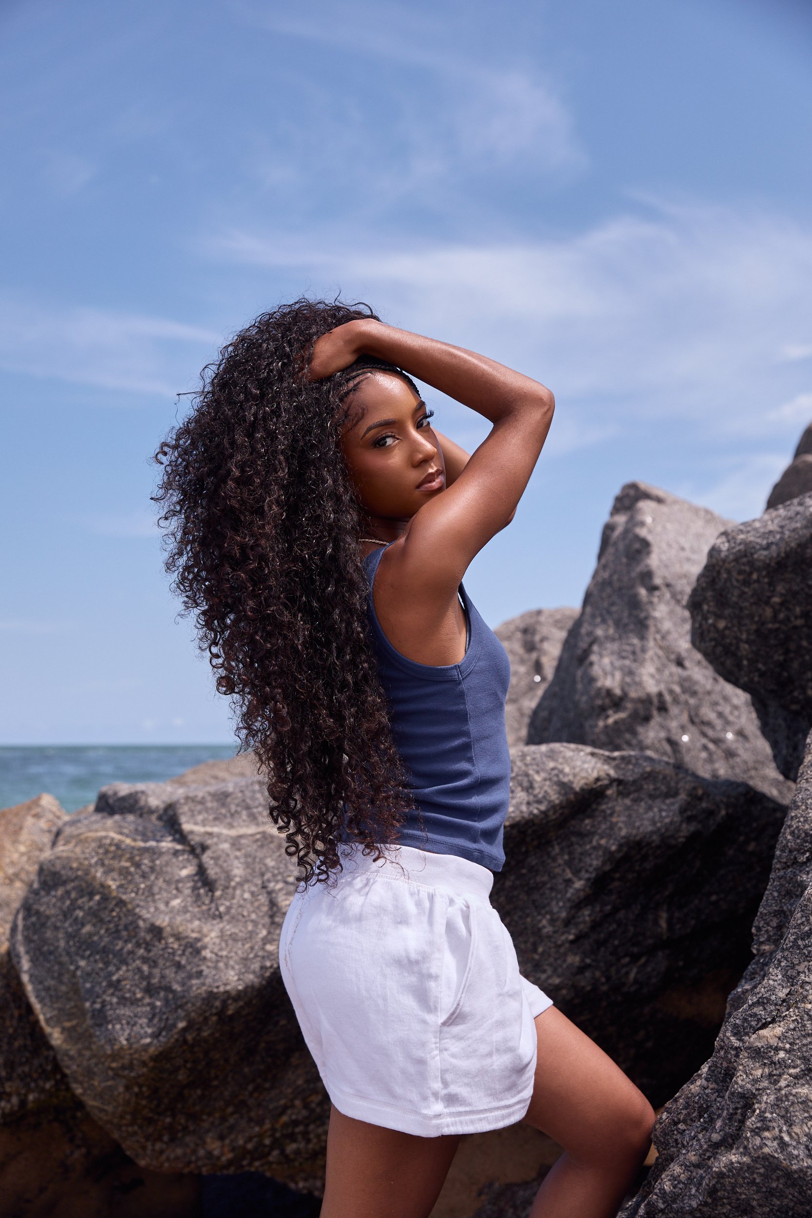 A woman with long curly hair standing among large rocks on a beach, wearing a blue sleeveless top and white shorts, with a partly cloudy sky in the background.