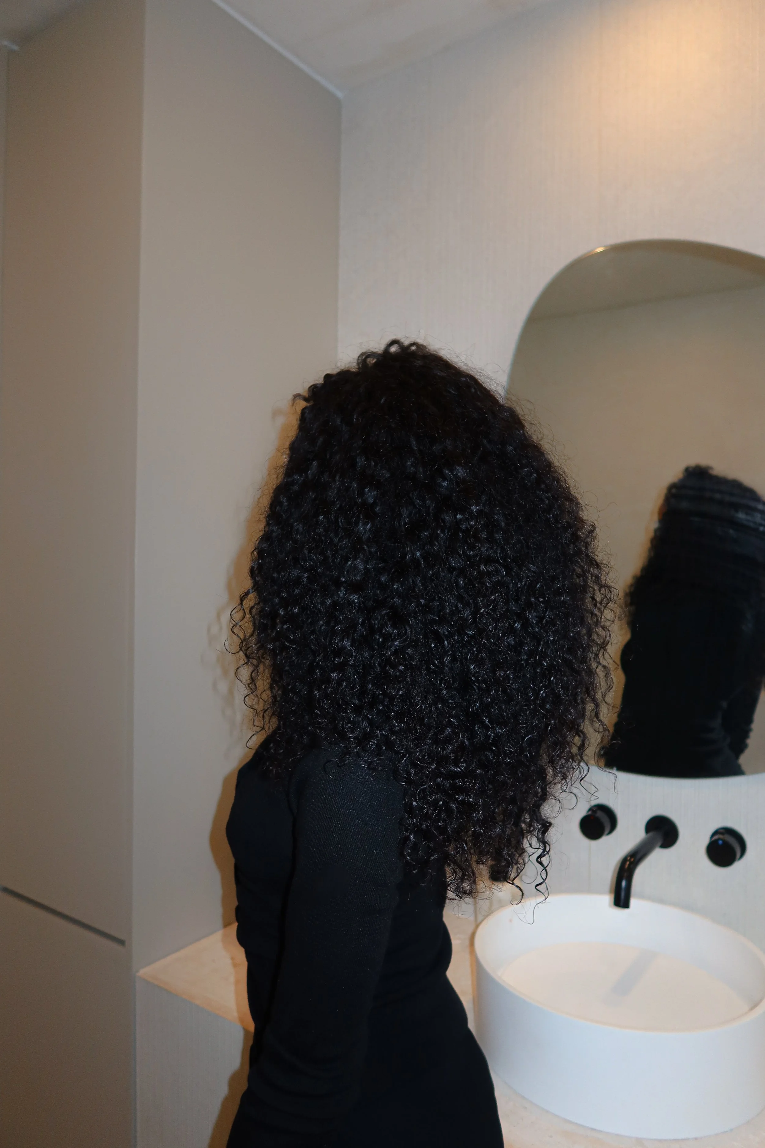 Back view of a woman with long, curly black hair wearing a black top, standing in front of a mirror in a bathroom with a white sink and black faucet, beige walls, and a light-colored countertop.