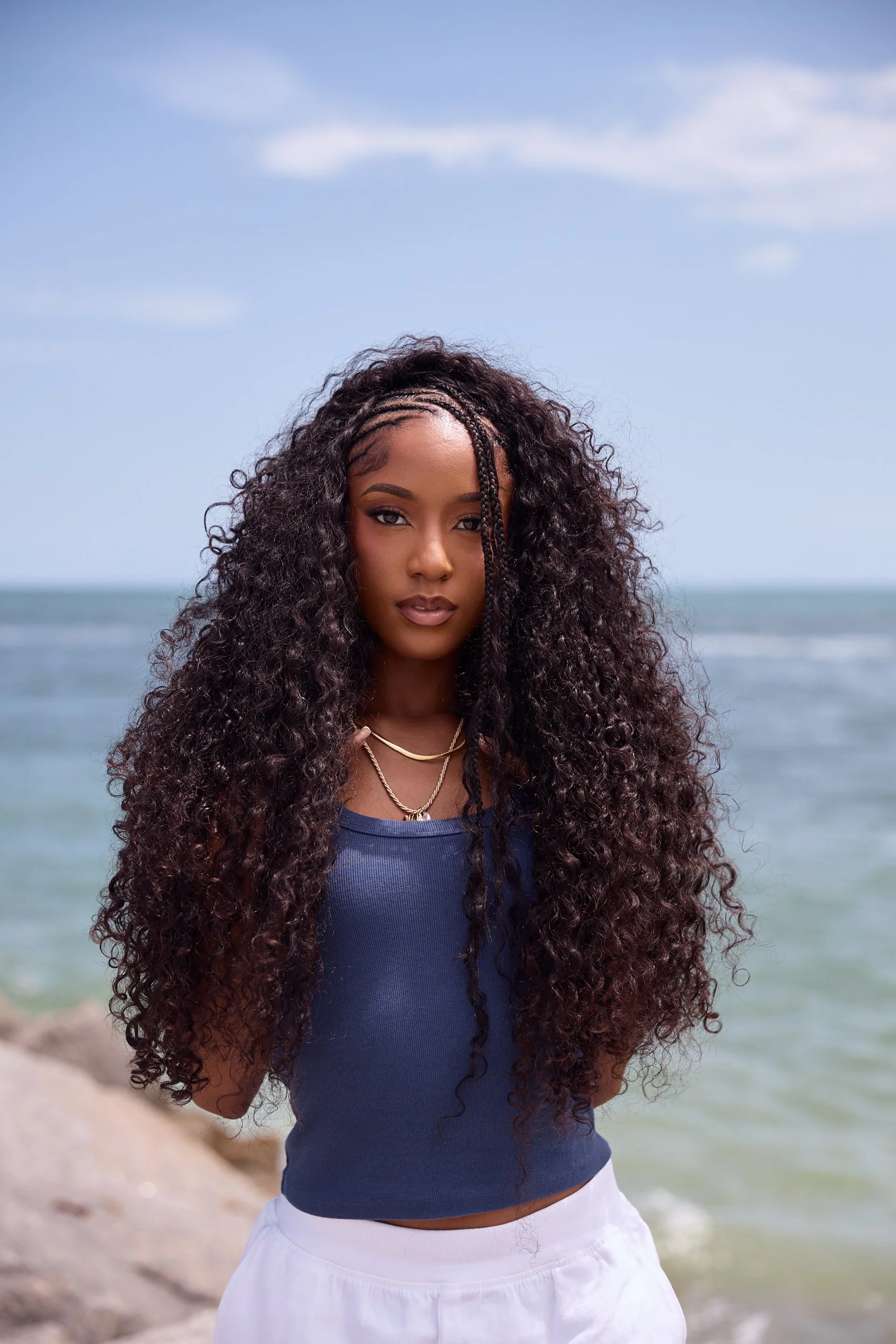 A woman at the beach with long curly hair, wearing a blue top and white shorts, standing in front of the ocean with a blue sky and some clouds.