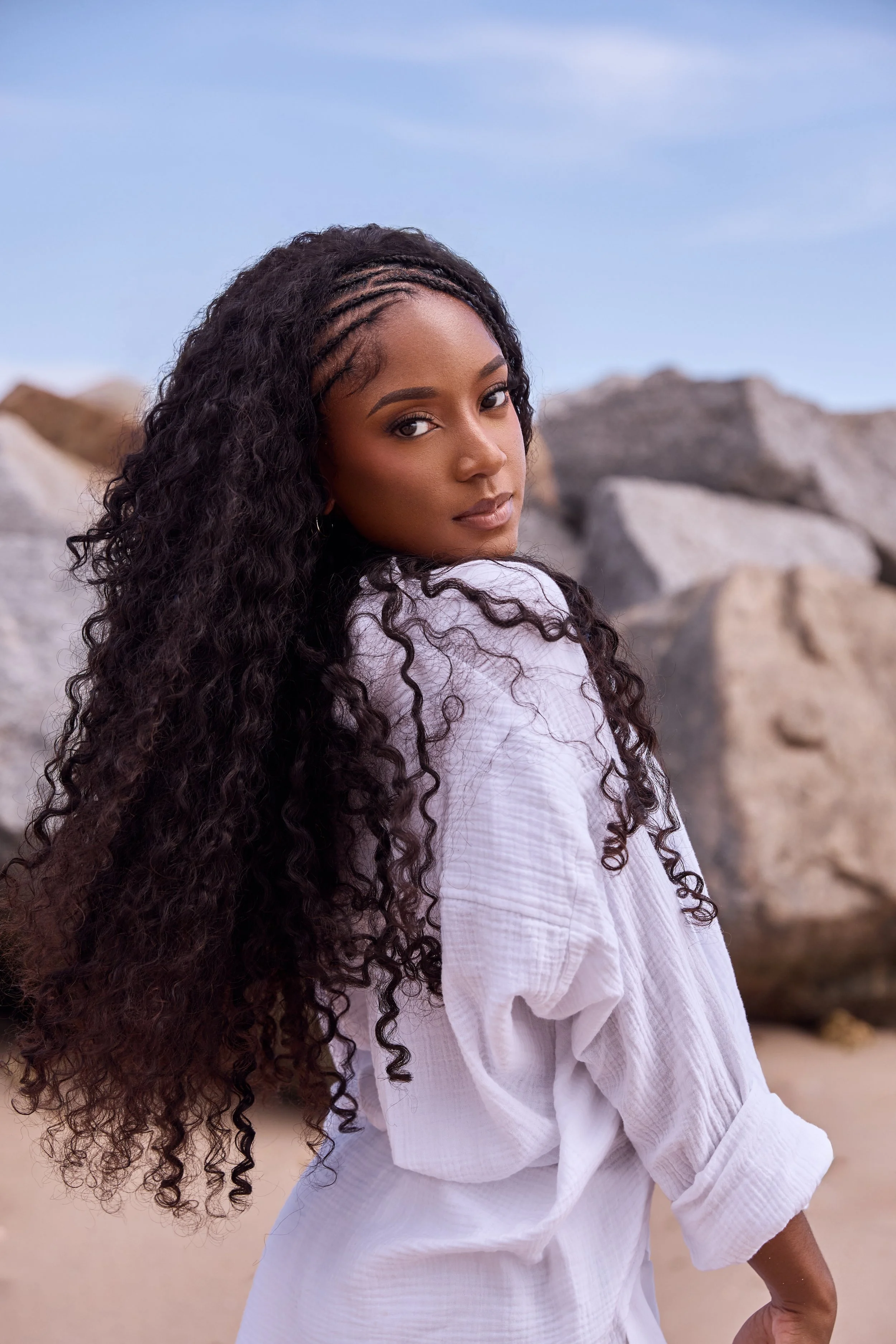 A woman with long, curly black hair wearing a white, textured, long-sleeve dress standing outdoors against a rocky background and a blue sky.