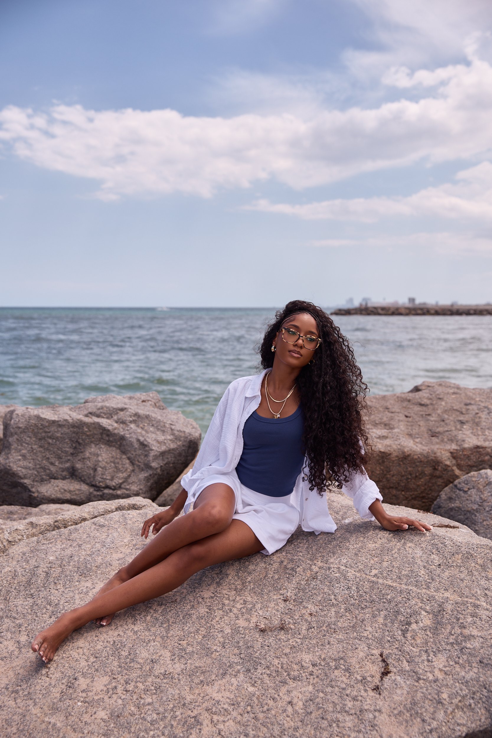 A woman with long curly hair, wearing glasses, jewelry, a blue top, and a white shirt, sitting barefoot on large rocks by the ocean with a cloudy sky overhead.