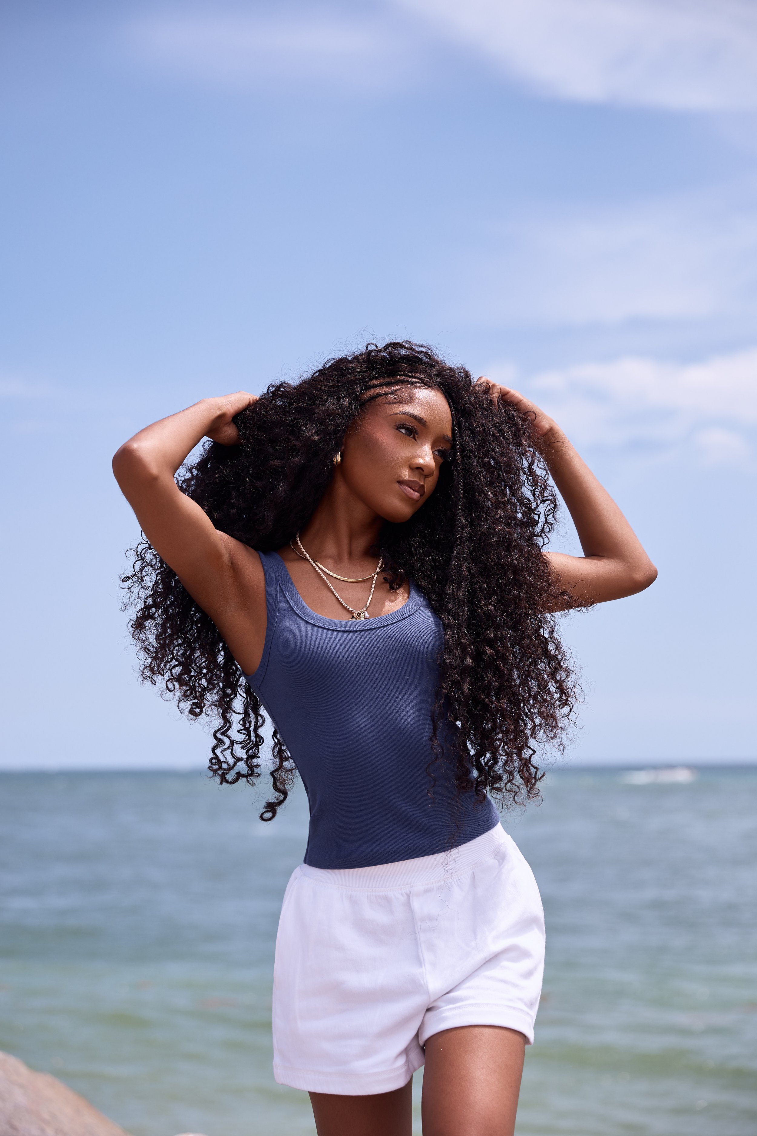 Young woman with curly hair standing on beach, wearing a blue sleeveless top and white shorts, with ocean and sky in background.