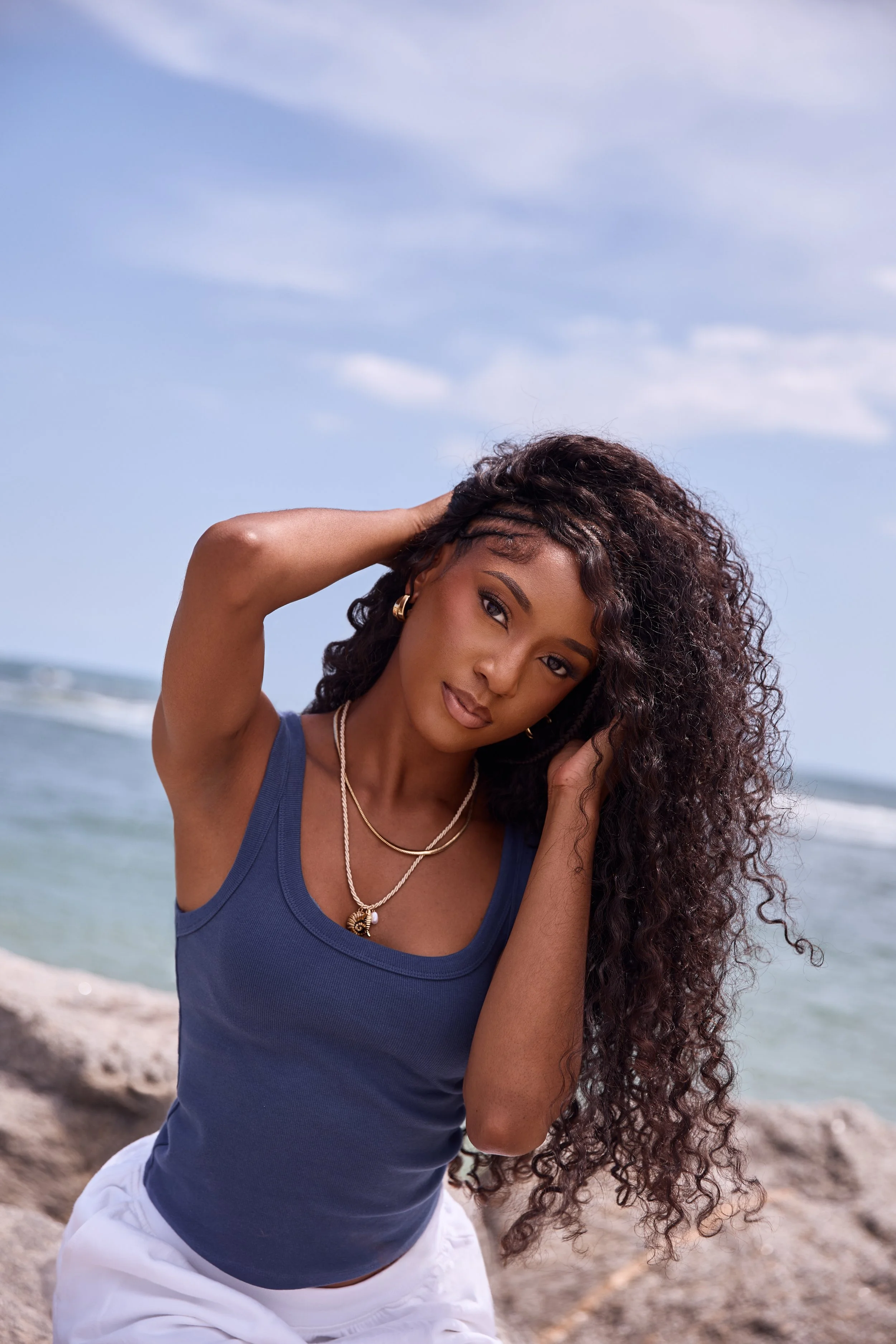 Woman with long curly hair wearing a blue tank top and white pants, at the beach with rocks and ocean in the background, holding her hair with one hand and wearing jewelry.