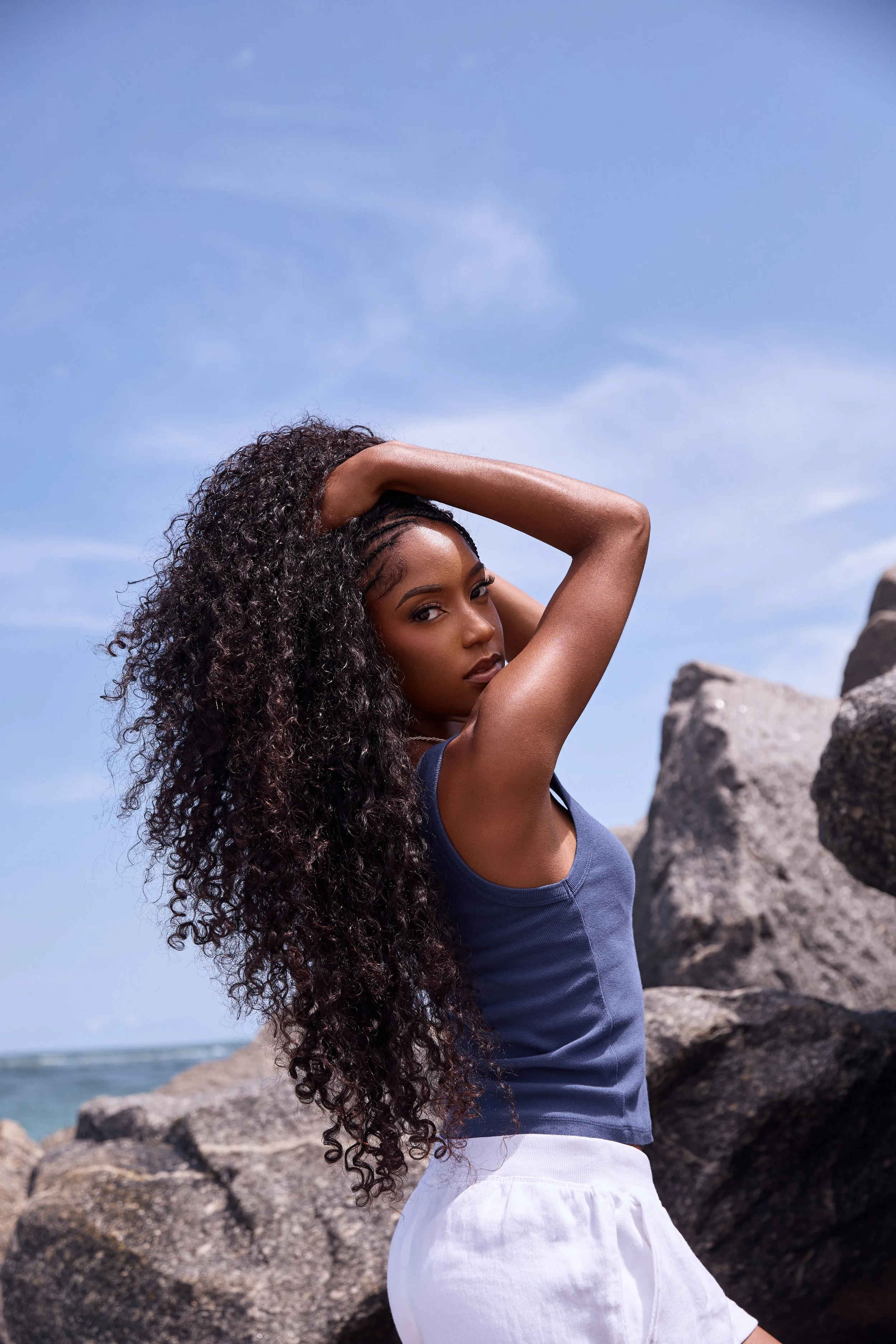 A woman with long curly hair standing on a rocky beach, with the ocean and a blue sky in the background, wearing a navy tank top and white shorts.