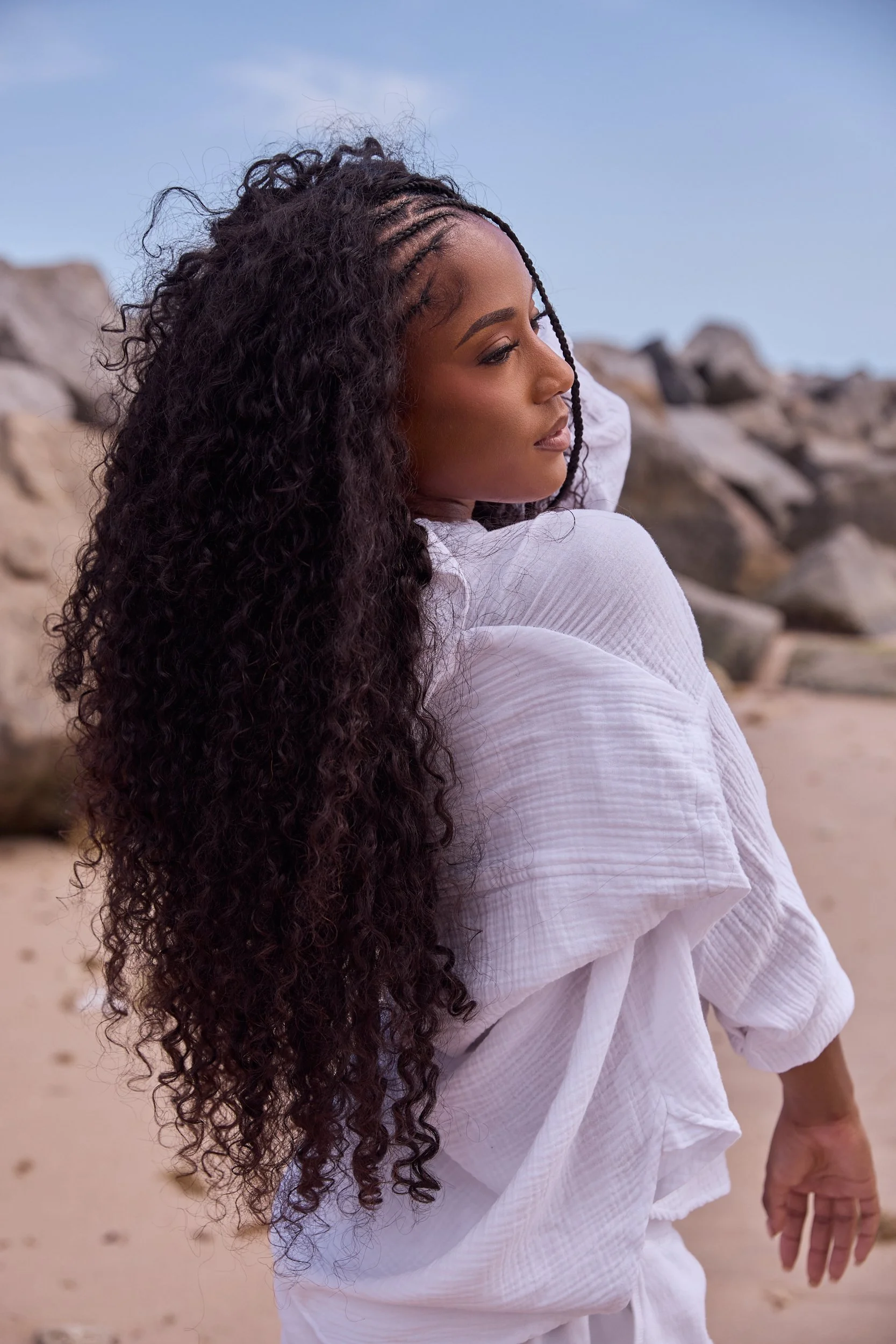 A woman with long curly hair and braided section, wearing a white shirt or dress, standing on a beach near rocks, with a blue sky overhead.