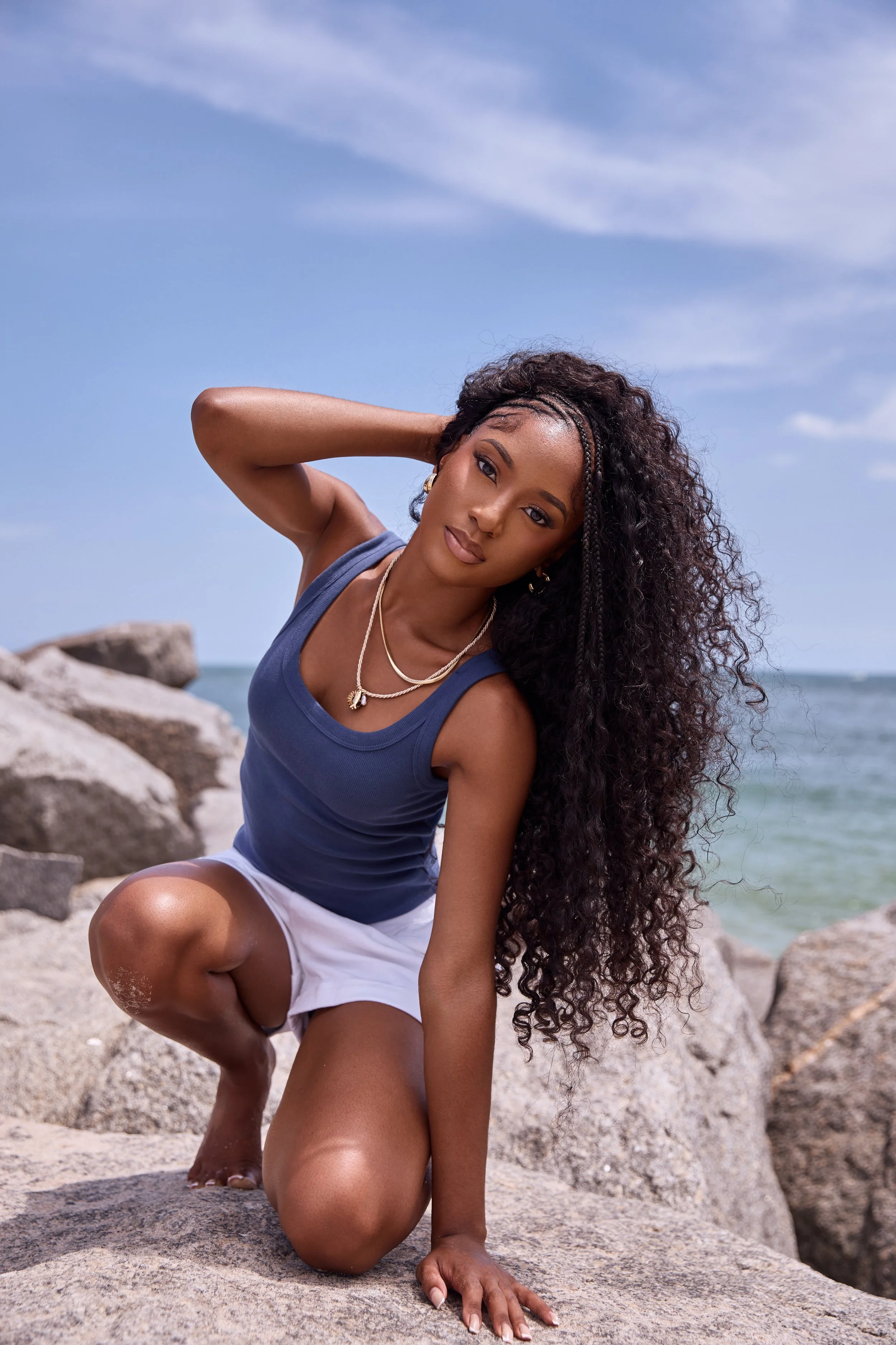 A woman with long, curly hair in a blue tank top and white shorts poses on rocks at the beach, with the ocean and sky in the background.