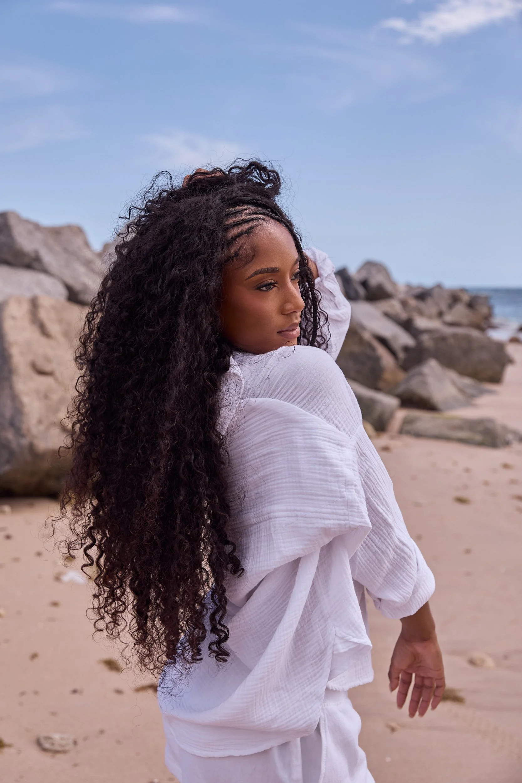 A woman with long curly hair in a white outfit standing on a beach with rocks and the ocean in the background.