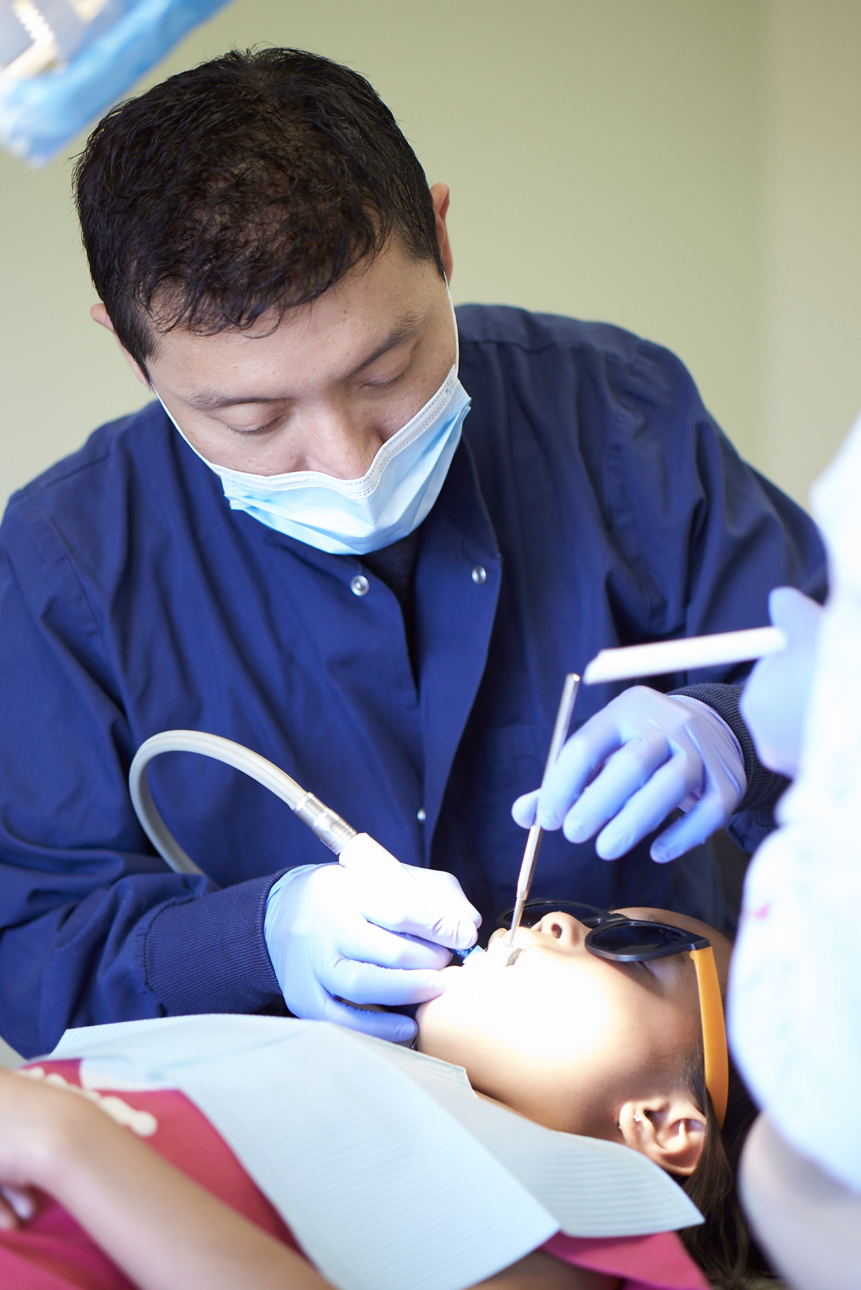A dentist in blue scrubs and a face mask provides dental treatment to a young girl lying in the dental chair, wearing protective glasses.