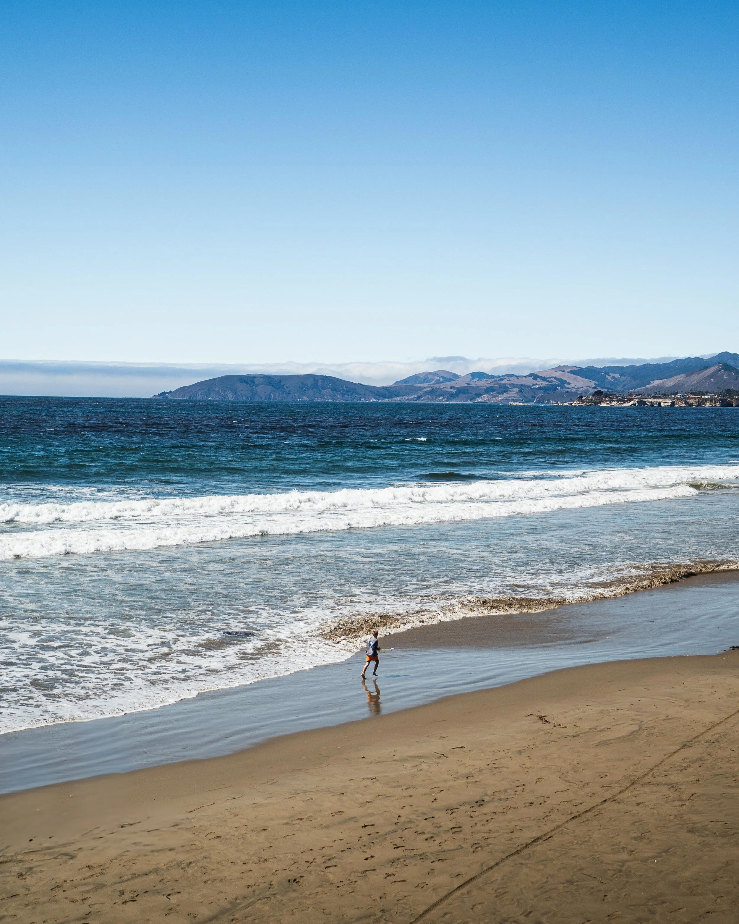 A person running along the shoreline of a beach with ocean waves and distant landforms under a clear blue sky.