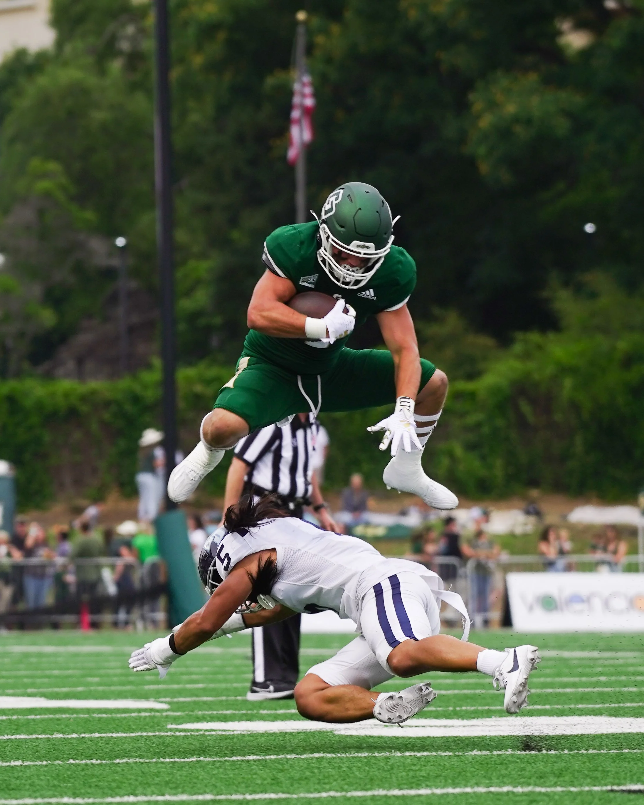 A football player in a green uniform jumps over a tackled opponent in a white uniform on a football field.