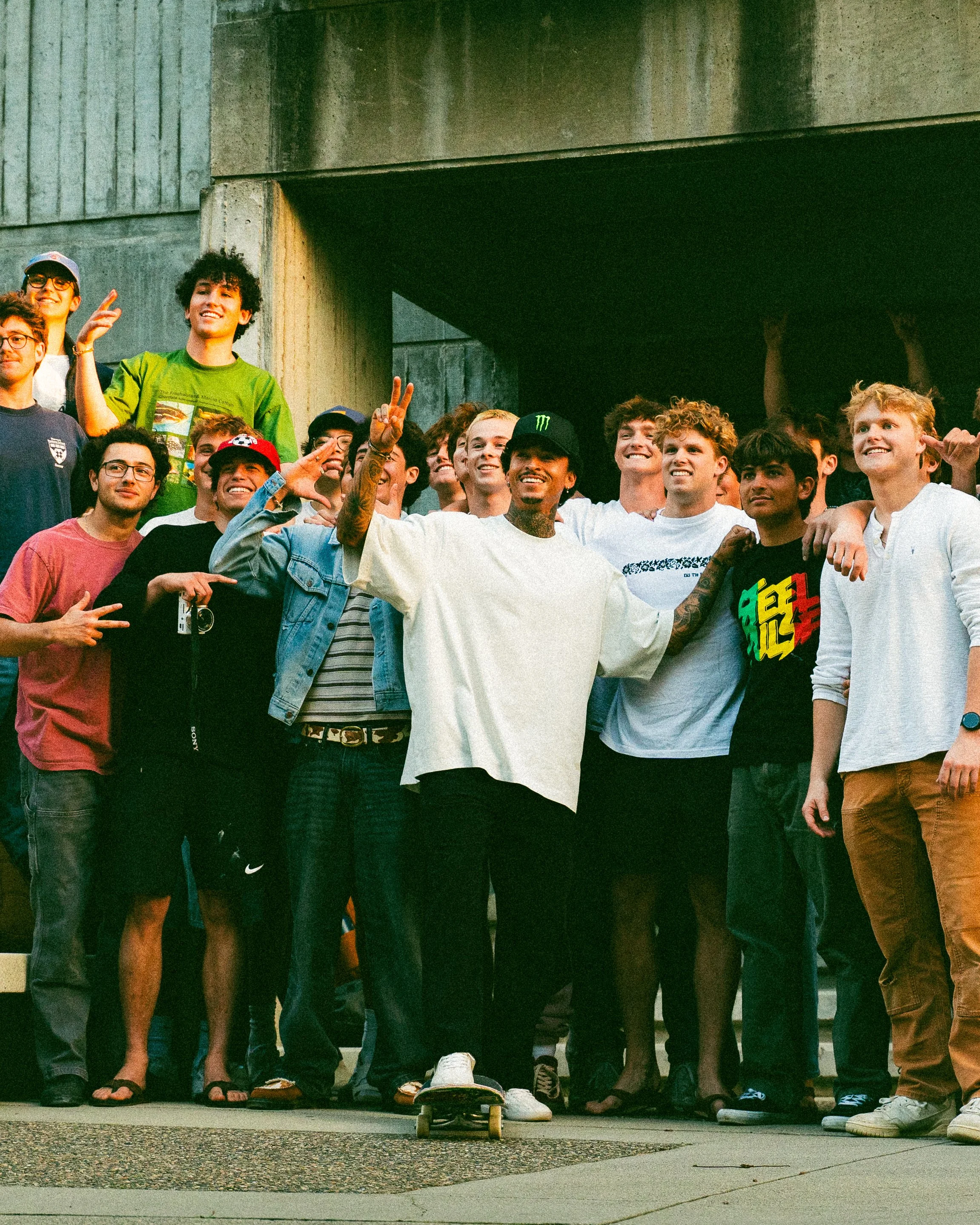 Group of young people and a man with tattoos, some holding up peace signs, standing under a concrete structure.
