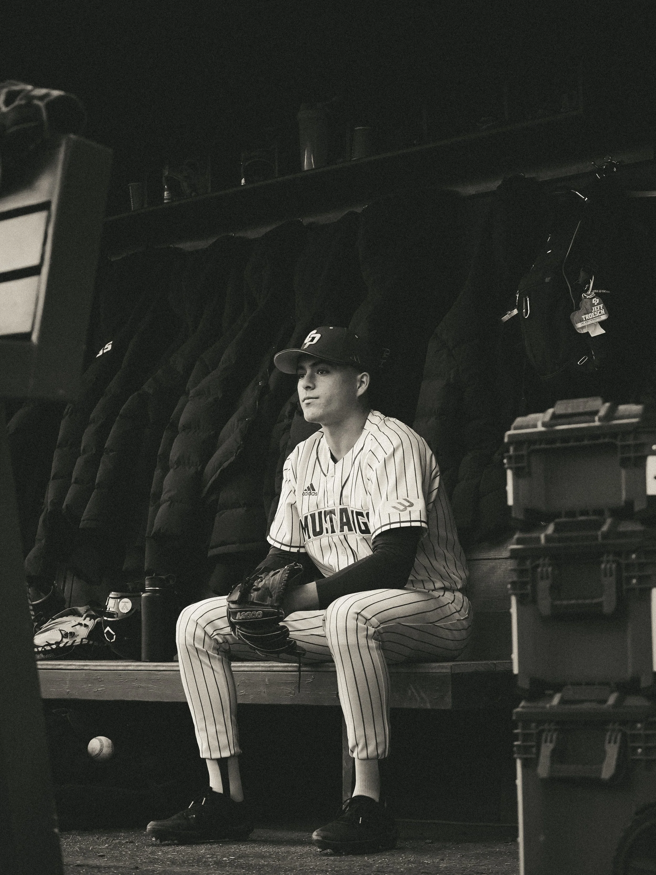 A young baseball player in a pinstripe uniform sitting on a bench in the dugout with a glove on his left hand, black cap, and cleats, against a background of jackets and equipment.
