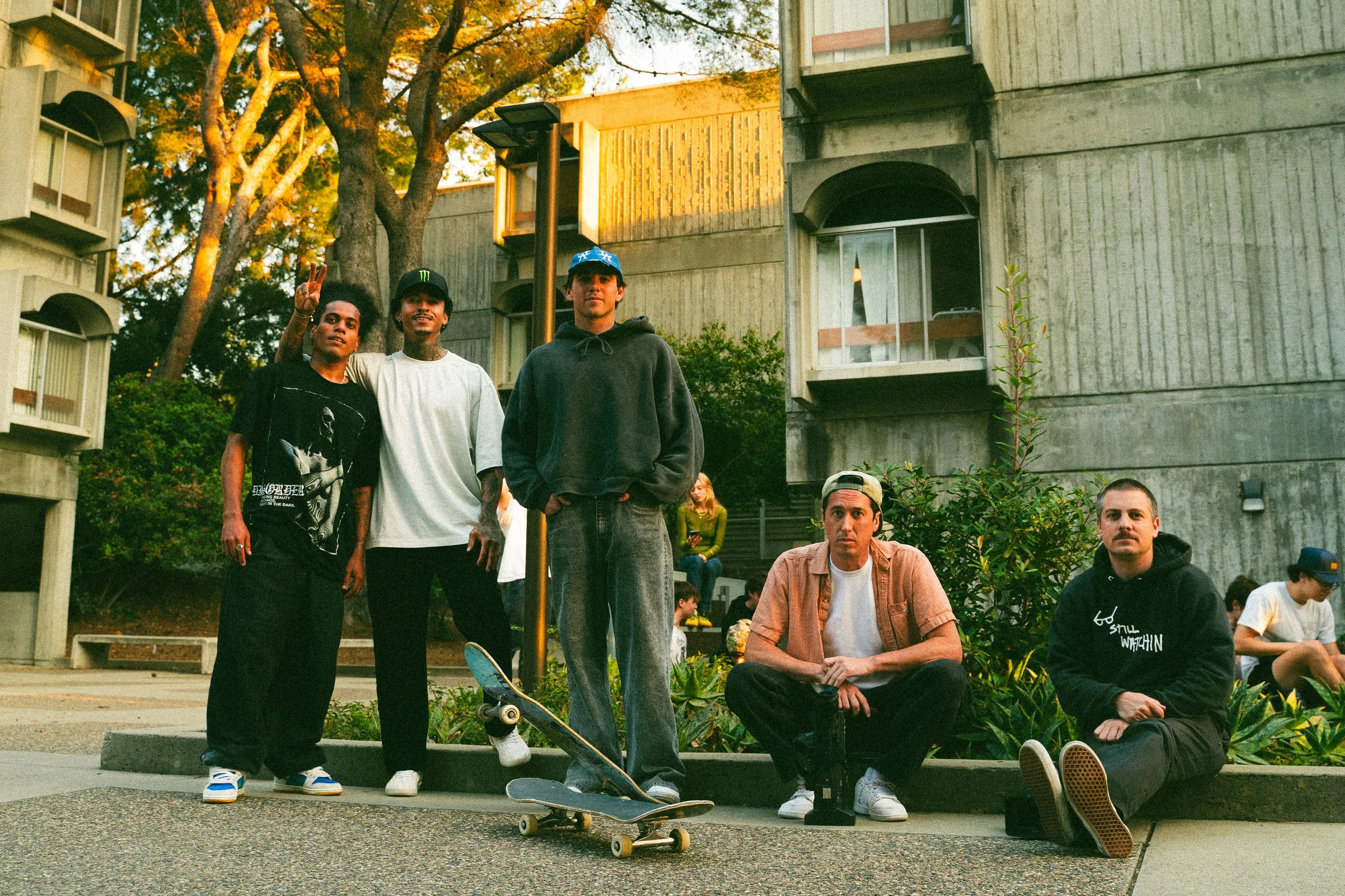 A group of five young men posing outdoors in front of apartment buildings with trees and bushes in the background. Some are standing, two are sitting on the ground, and one has a skateboard placed in front of him.