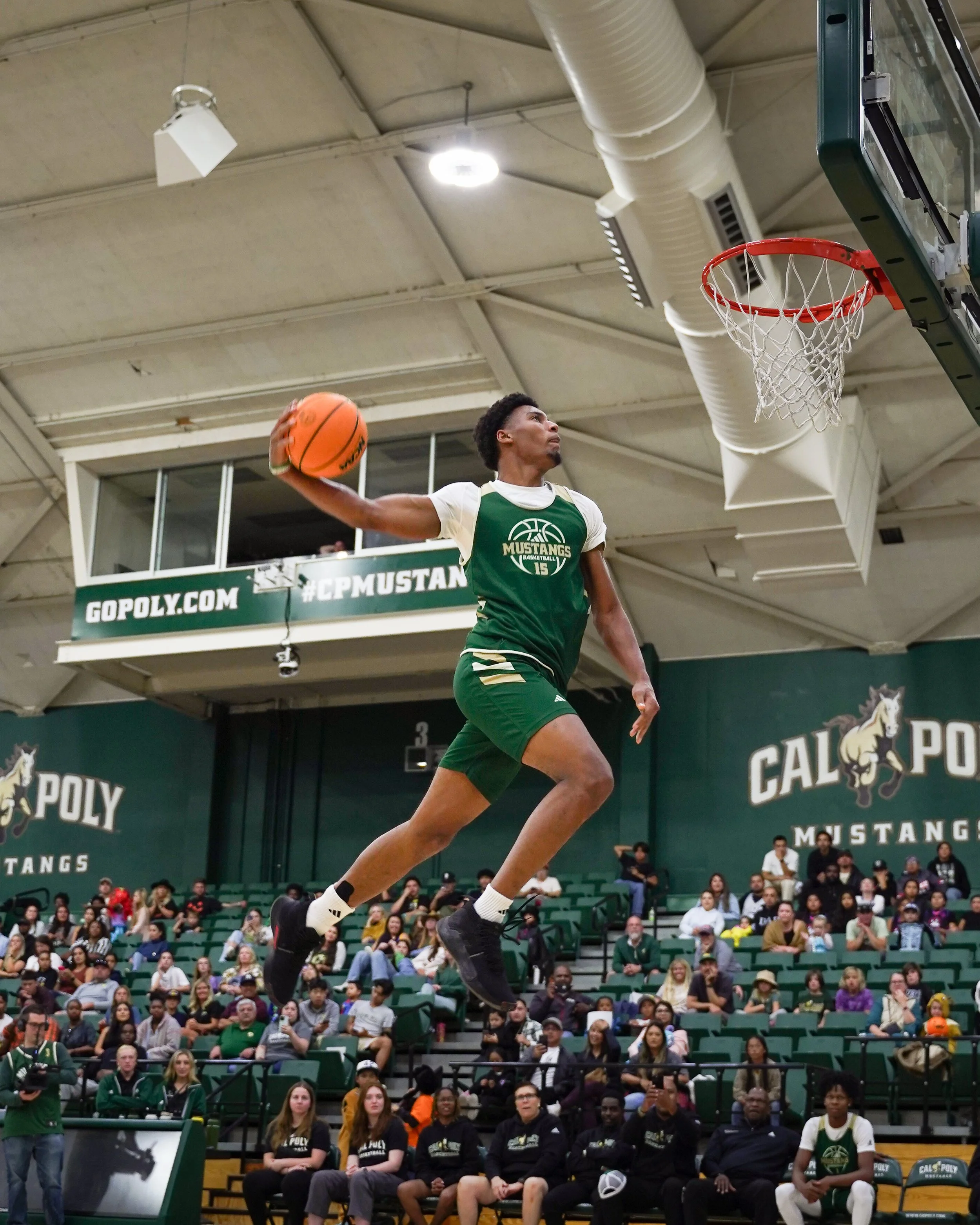 A male basketball player in green and white uniform jumping to make a shot during a game at Cal Poly Mustangs gym.