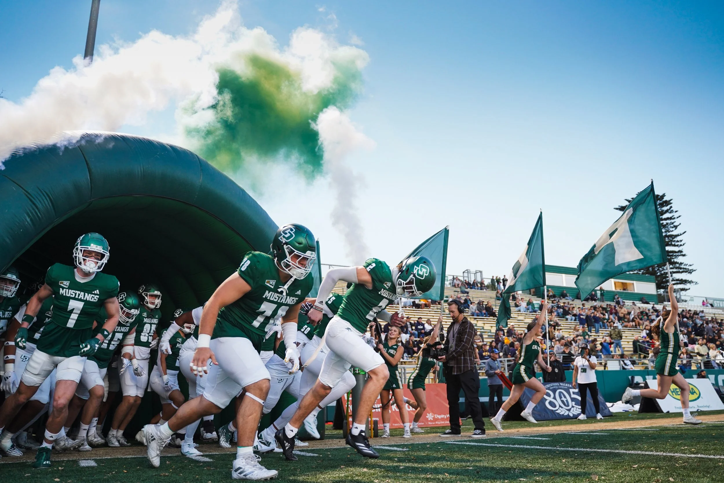 San Diego State Aztecs football team entering the field through a tunnel with green smoke and cheerleaders waving flags, on a sunny day with spectators in the stands.