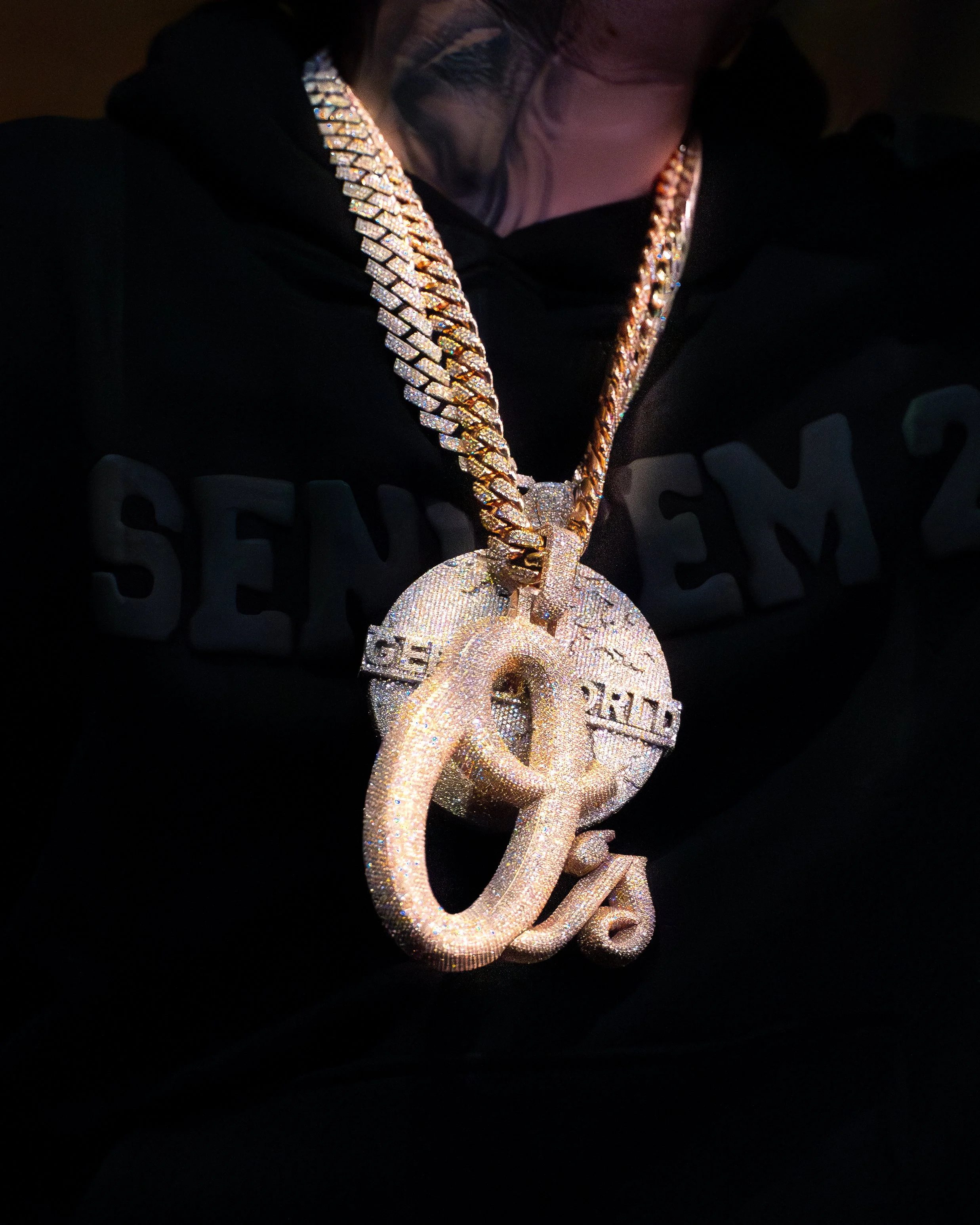 Person wearing a black hoodie with a large gold chain and a pendant featuring the percentage symbol (%) and the words "Keep It Real" encrusted with diamonds.