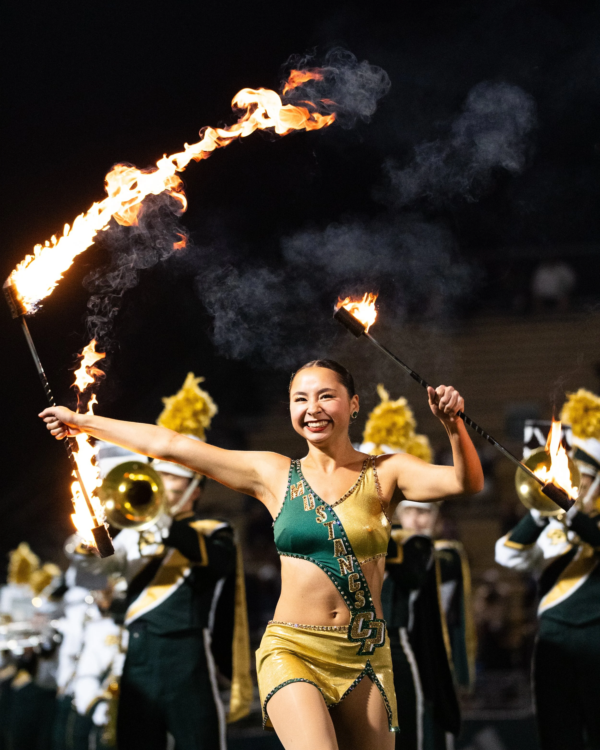 A cheerleader in a green and gold costume with 'MSTAN' and 'G' on it, holding fire torches during a performance, with a marching band in the background.