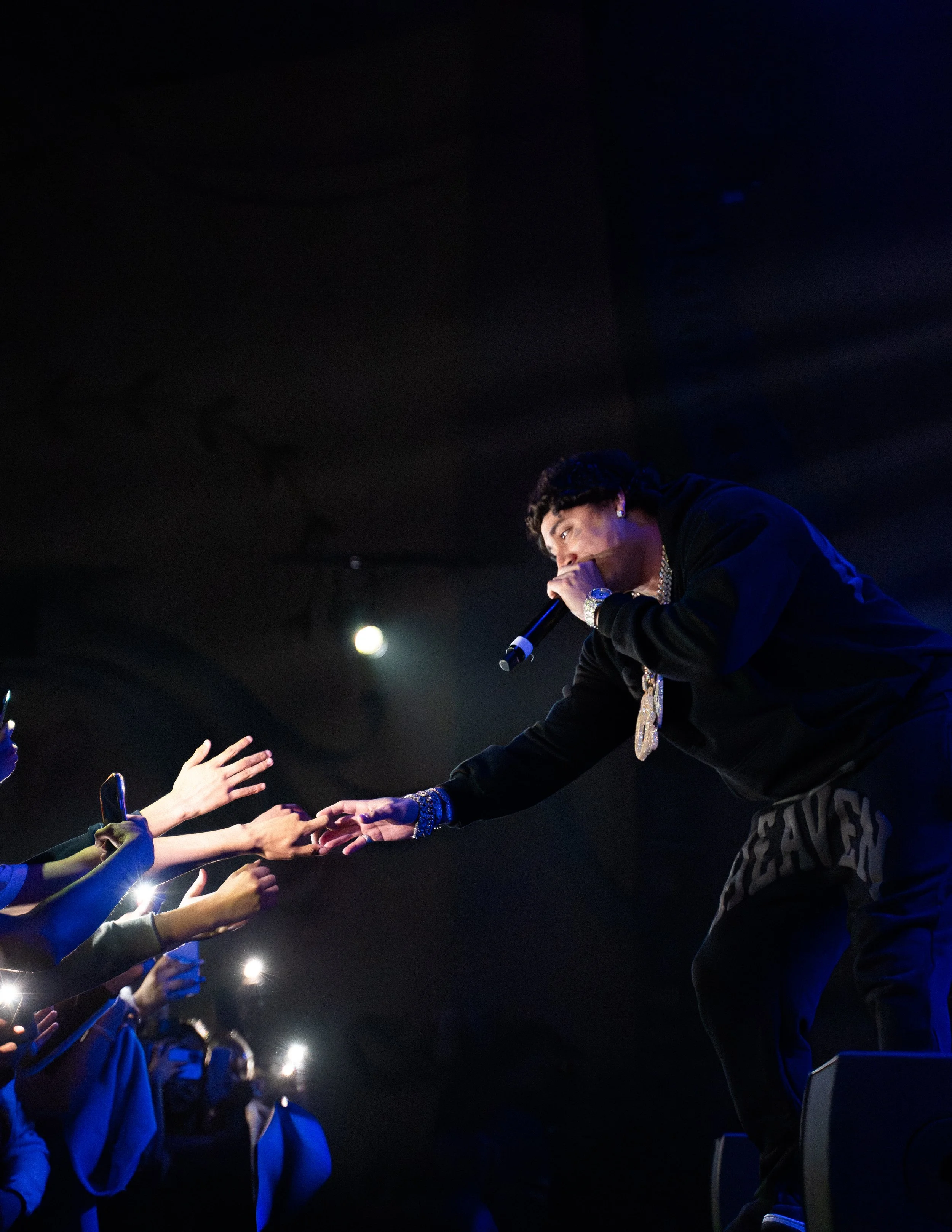 Male performer reaching out to audience during concert, fans with smartphones taking pictures.
