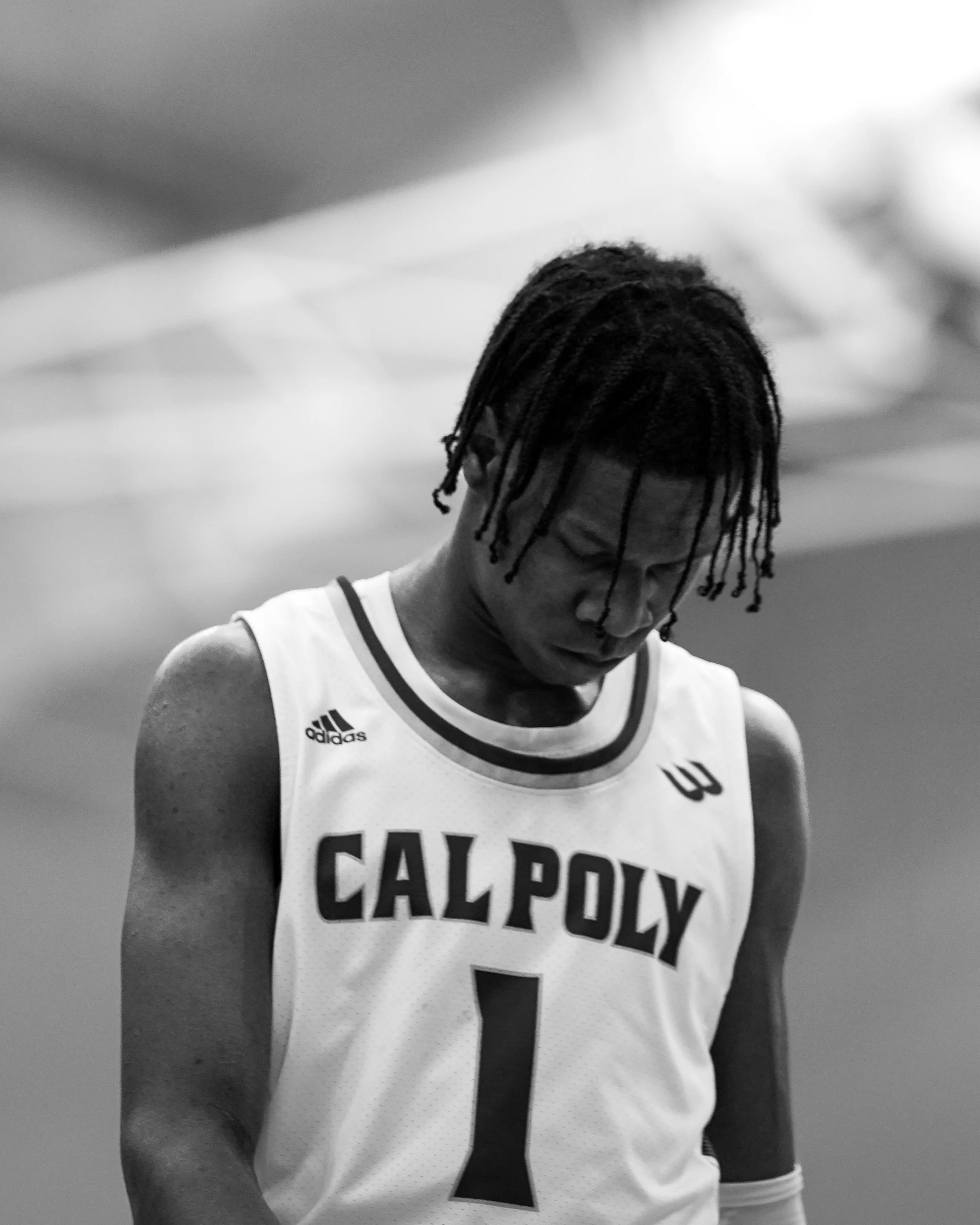 Black and white photo of a basketball player wearing a jersey with 'CALPOLY' and the number 1, looking down in a contemplative pose.
