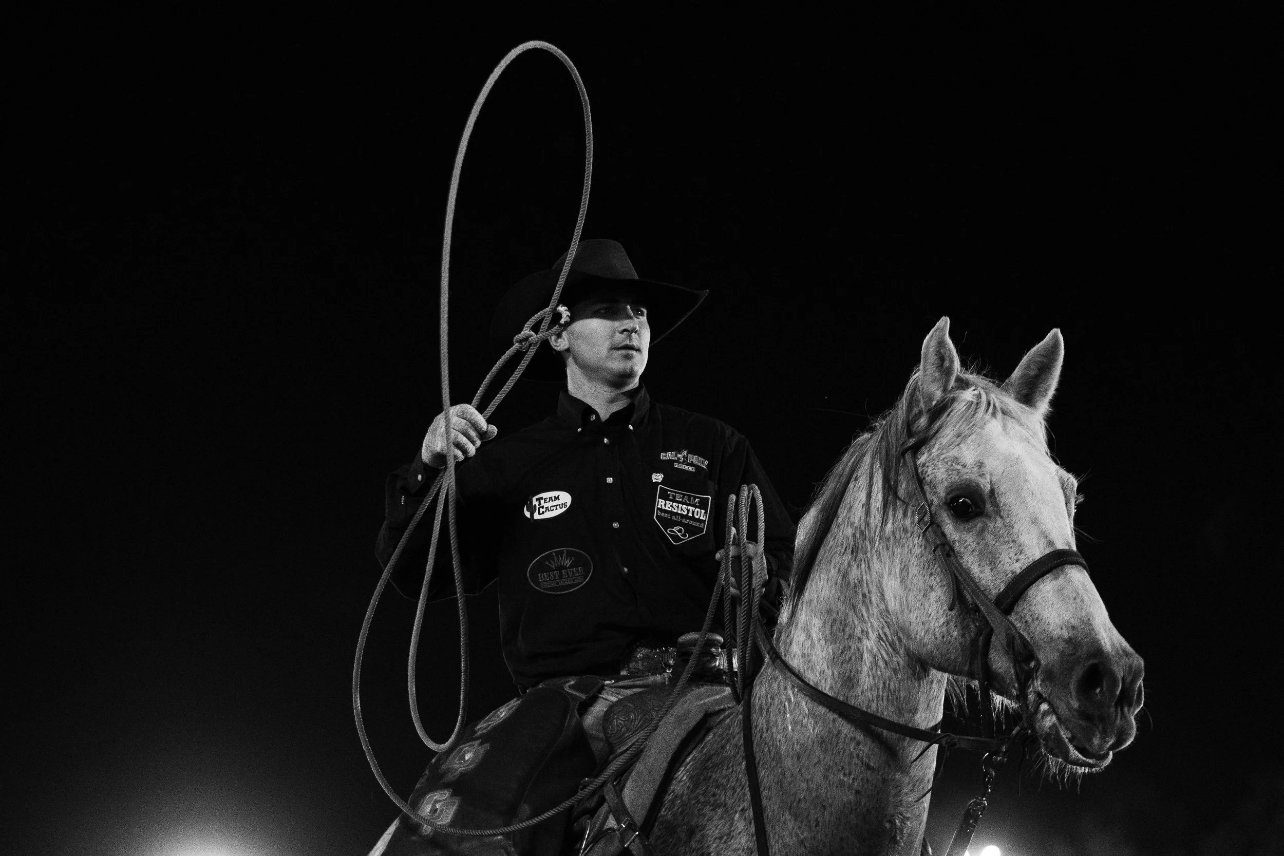 A man on horseback wearing a black cowboy hat and shirt, holding a rope, is captured in a black and white photo at night.