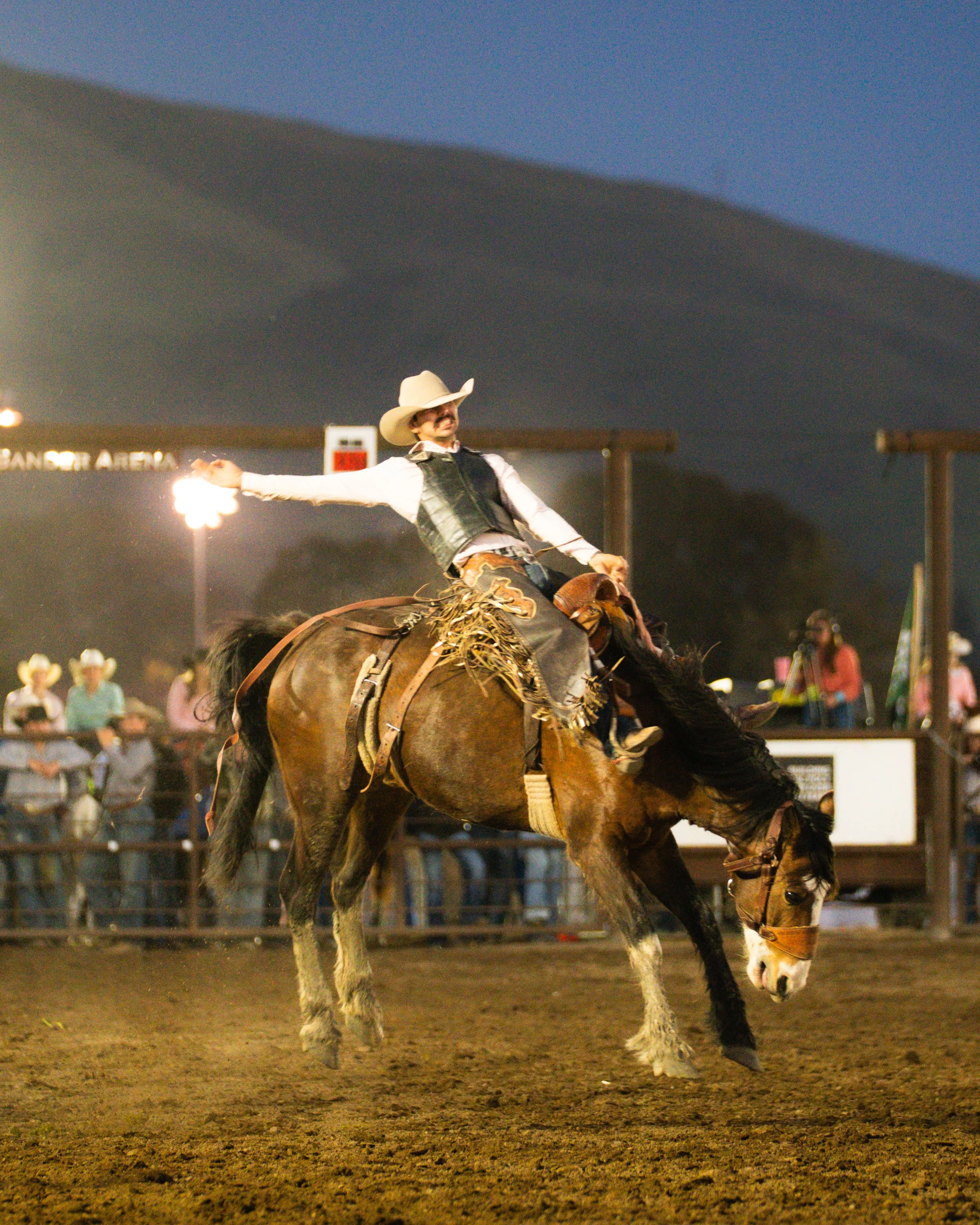A cowboy in a hat, vest, and Western attire riding a bucking horse at a rodeo event during the evening.