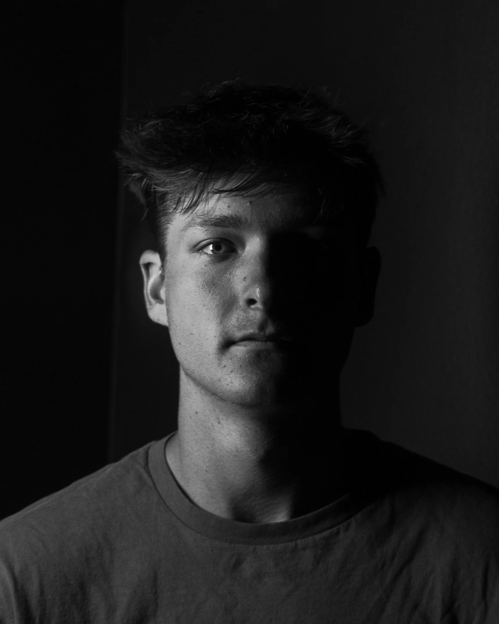 Black and white portrait of a young man with short hair, facing forward, with one side of his face illuminated and the other in shadow.
