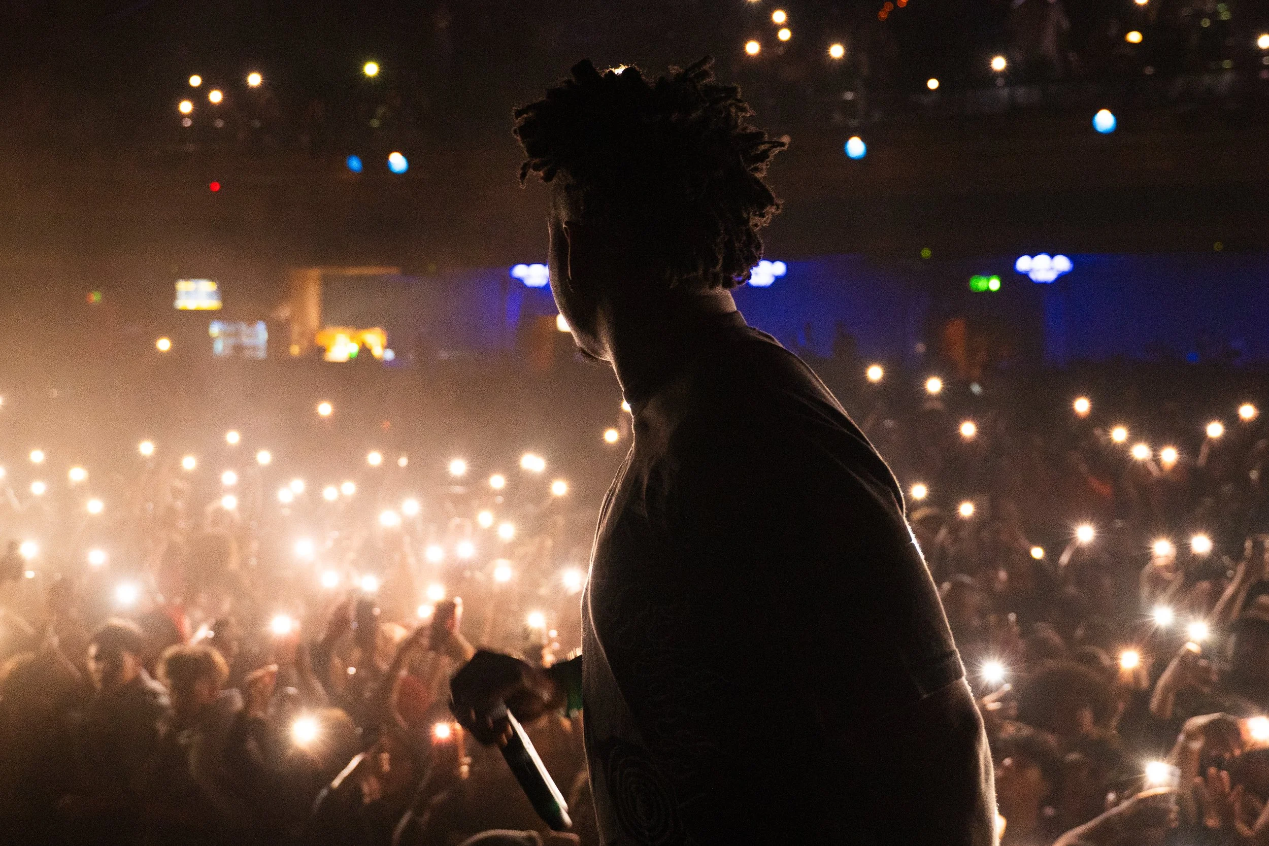 A performer with a bracelet on their wrist stands in front of a large crowd at a concert, with many audience members holding up their phones with flashlight on, creating a sea of lights.