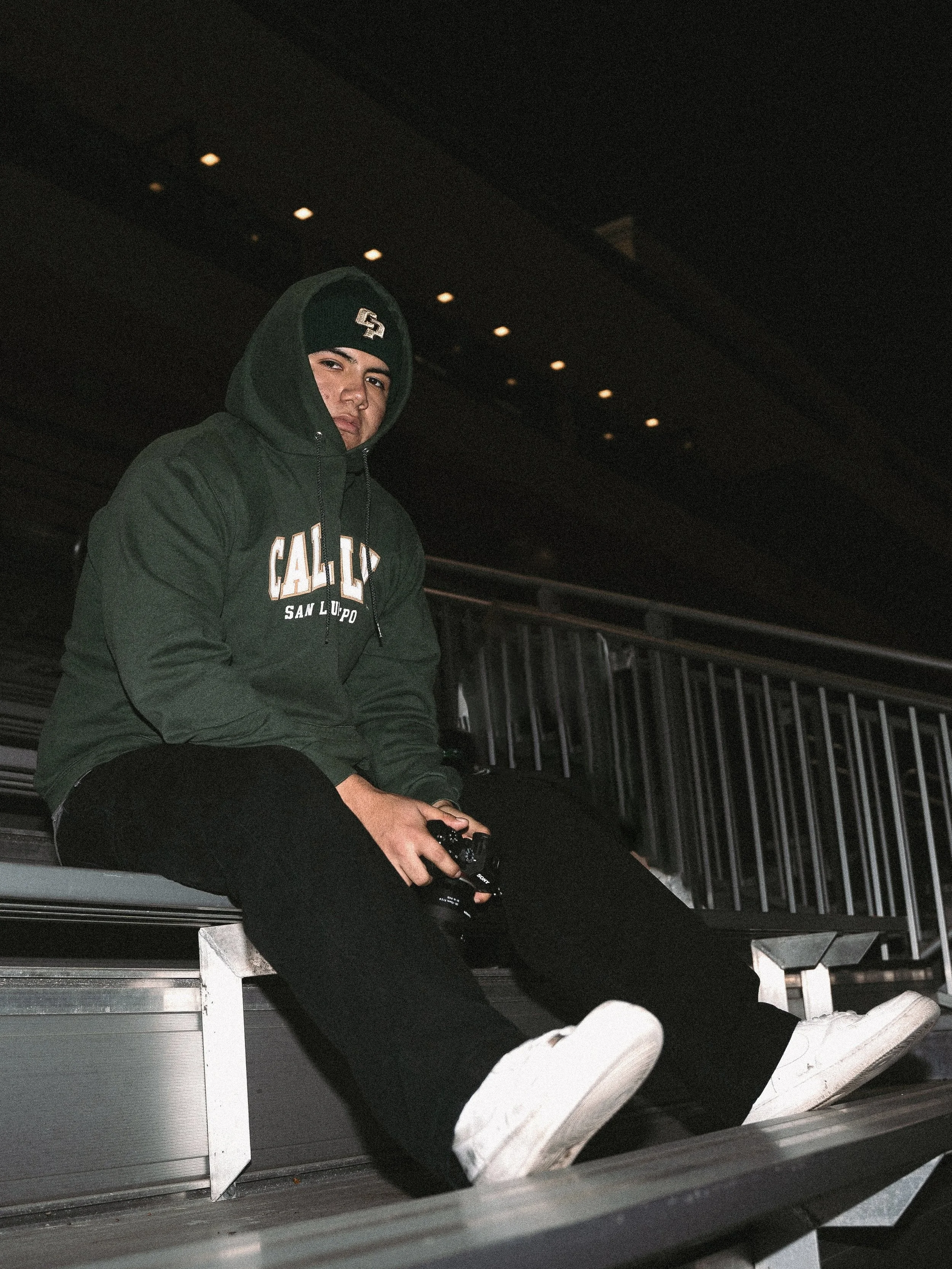 A young man in a green hoodie, black pants, and white sneakers sitting on metal bleachers in a dimly lit indoor setting, holding a camera in his right hand, with a serious expression and one eye slightly squinted.