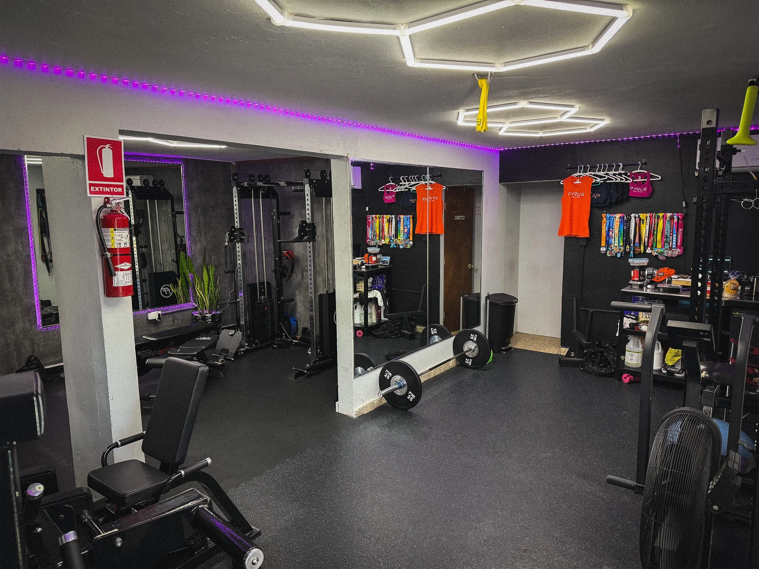 A gym with workout equipment, weights, and exercise machines. There are colorful lanyards hanging on a wall, shirts hanging on a rack, and there's a mirror with purple LED lights around the ceiling.