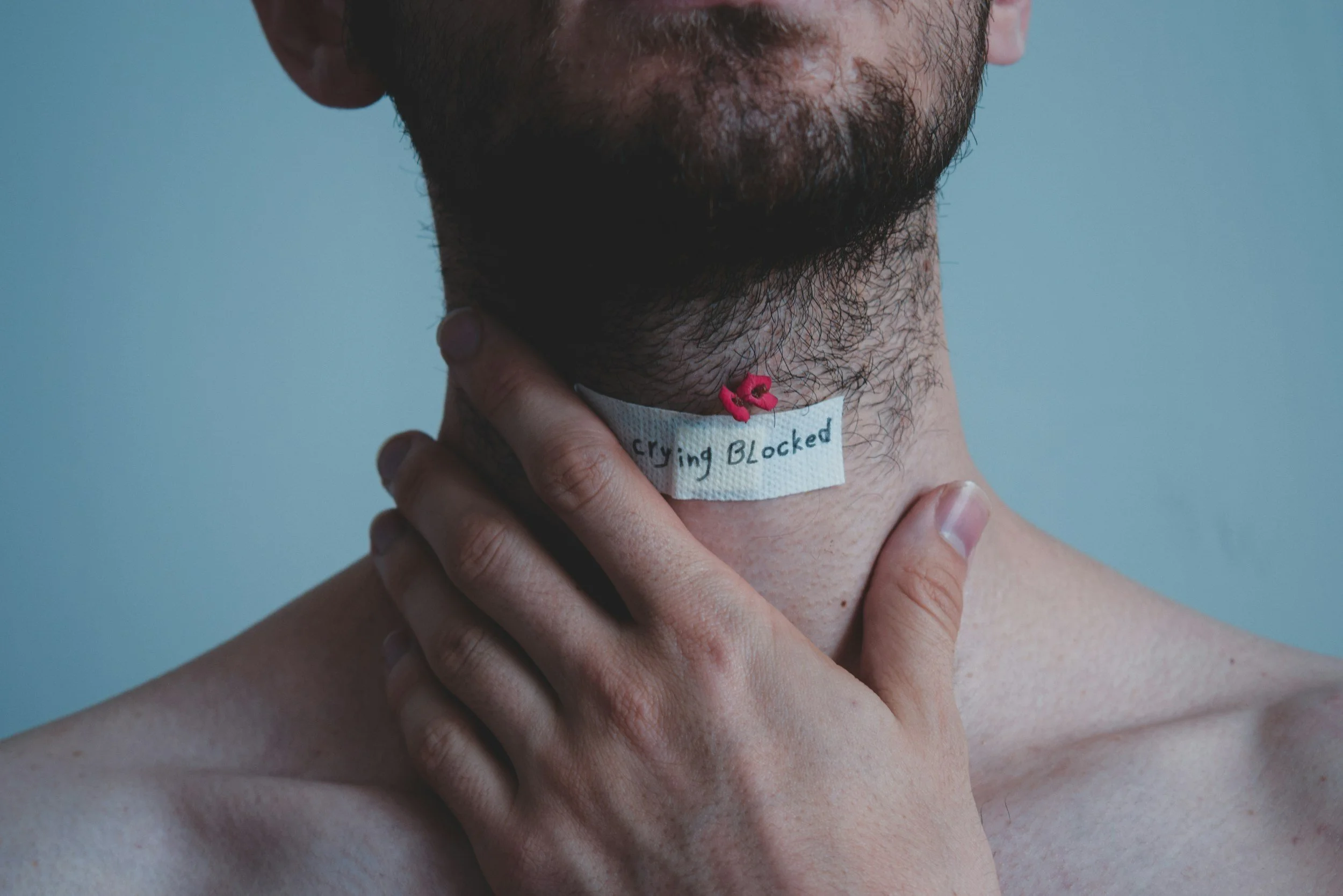 Close-up of a person with a beard holding their neck, showing a bandage with a handwritten message saying 'Crying Blocked' and a small pink object attached to it.