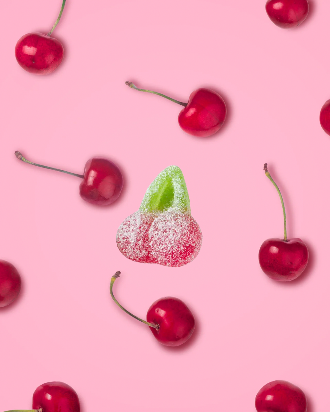 Gummy cherry and gummy heart candies placed on a pink background, surrounded by real cherries.