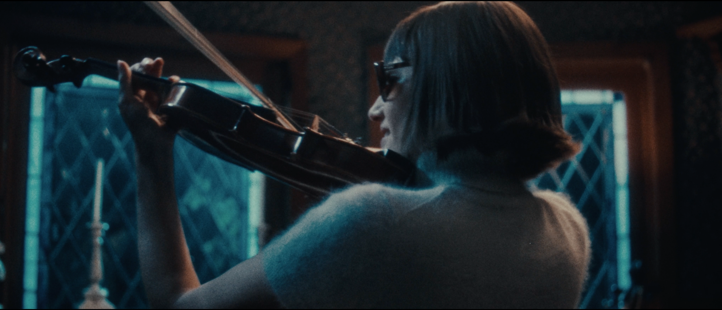 A woman with short hair and glasses playing the violin in a dimly lit room.