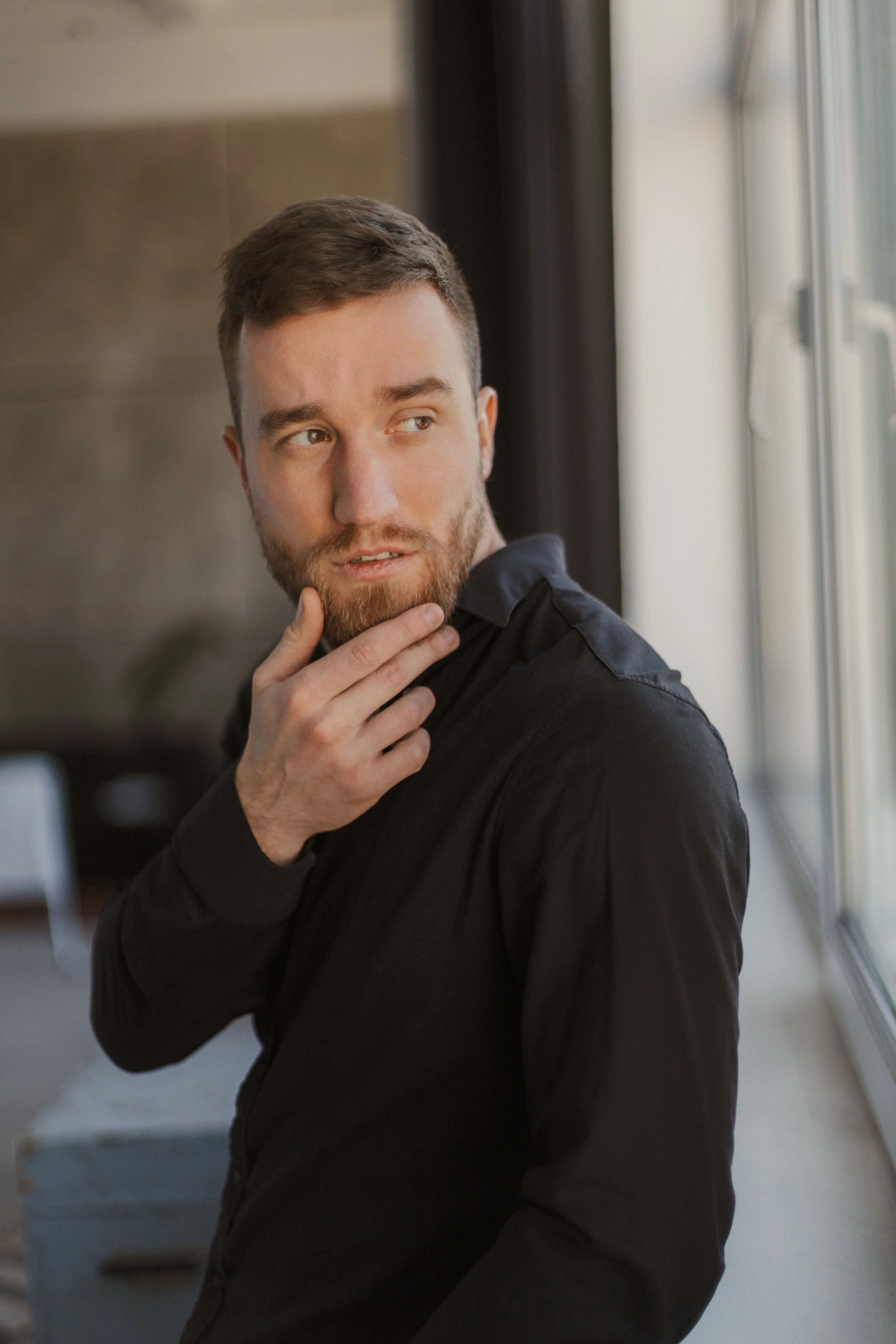 A man with short brown hair and a beard, wearing a black shirt, standing near a large window, looking thoughtful with his hand on his chin.