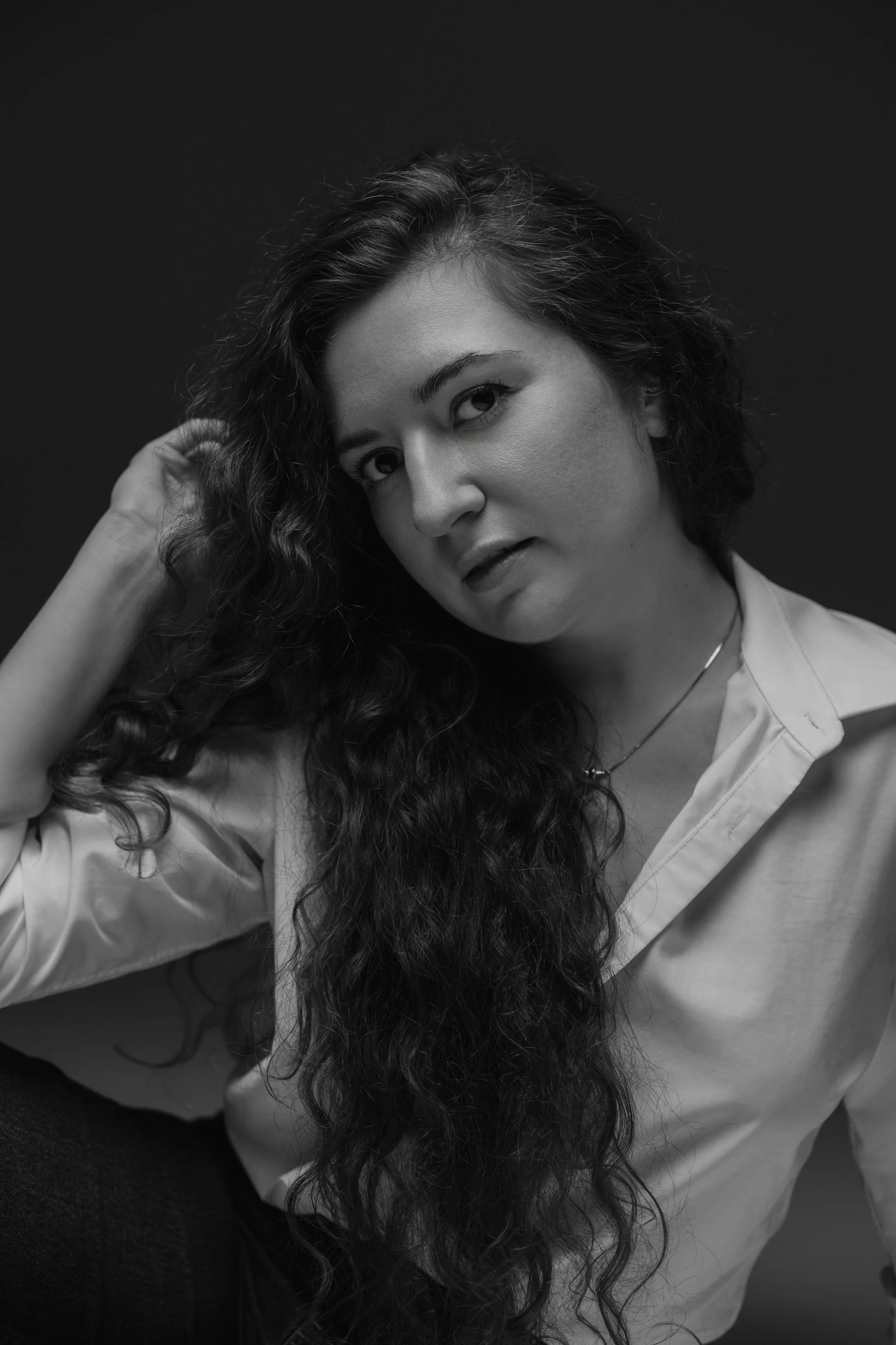 Black and white portrait of a woman with long curly hair, wearing a light-colored shirt and a necklace, looking at the camera with a slight tilt of her head.