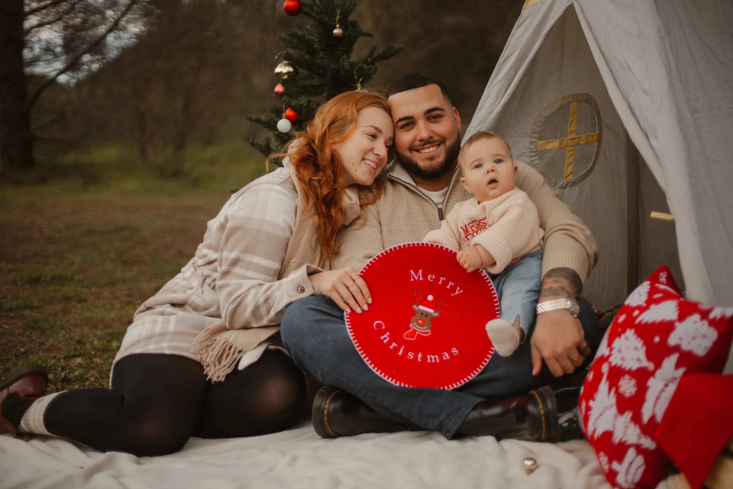 Family sitting on the ground outdoors next to a Christmas tree and a small tent, celebrating Christmas and holding a red plate with a reindeer and 'Merry Christmas' written on it.