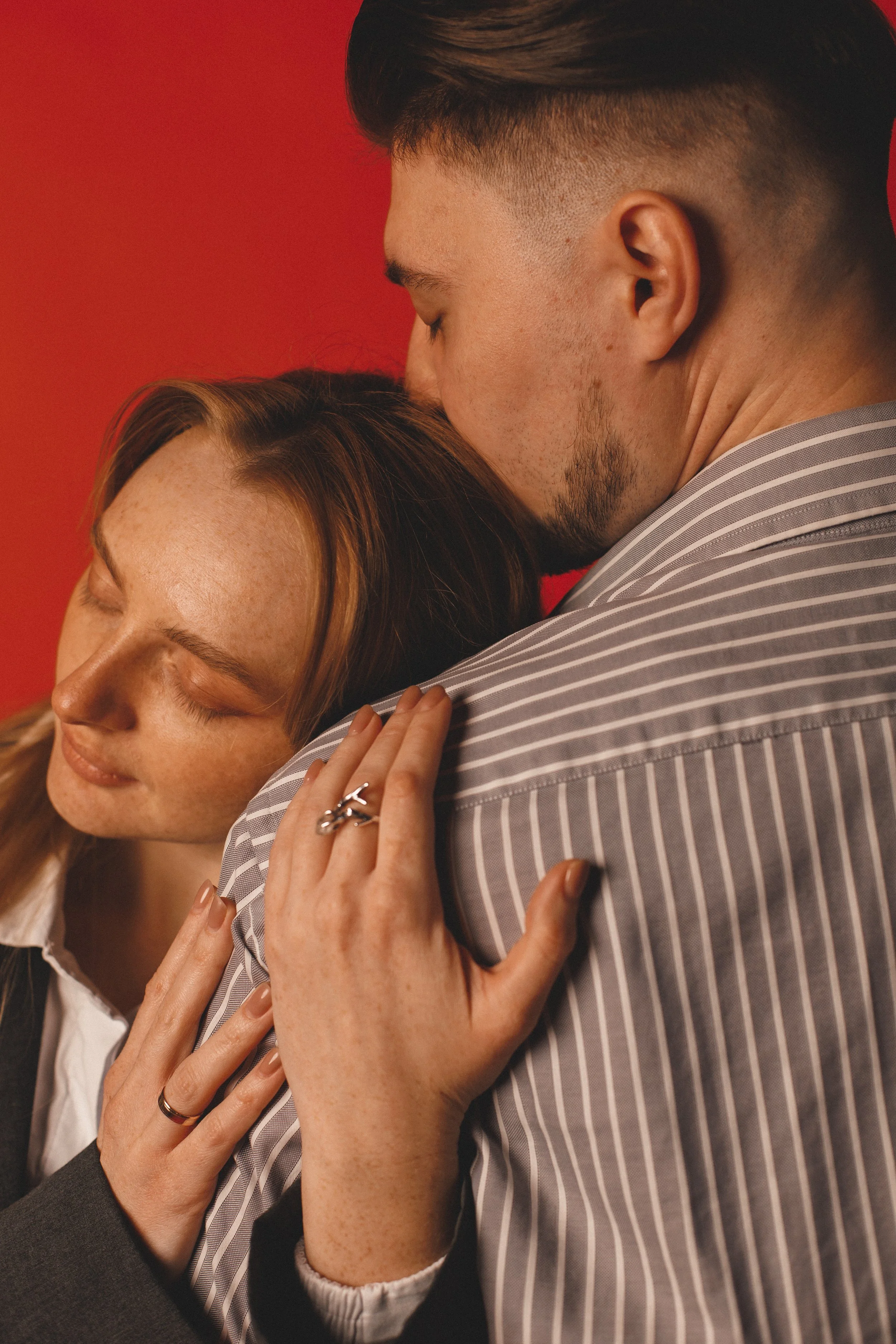 A man and woman share an intimate embrace, with the man kissing the top of the woman's head. The woman has closed eyes, light skin, freckles, and red hair, wearing rings on her fingers. The man has short dark hair, light skin, a beard, and is wearing