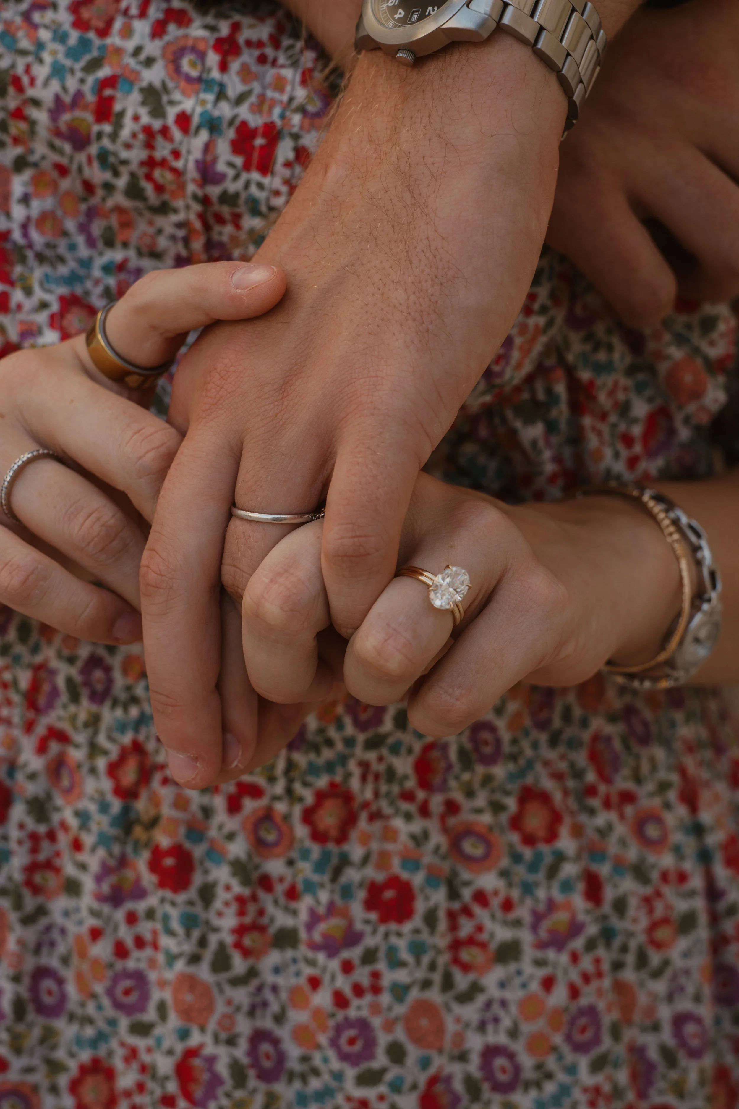 Close-up of four hands showing wedding rings, including a large diamond ring, against a floral patterned fabric background.