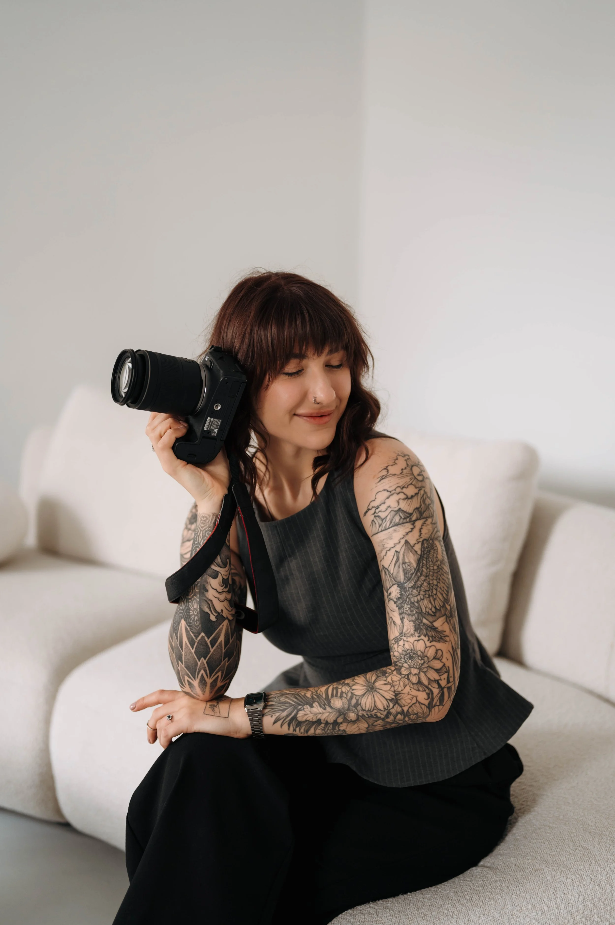 A woman with tattoos on her arms, dark hair, and a nose piercing, sitting on a light-colored sofa, holding a camera to her shoulder with her eyes closed and a slight smile.