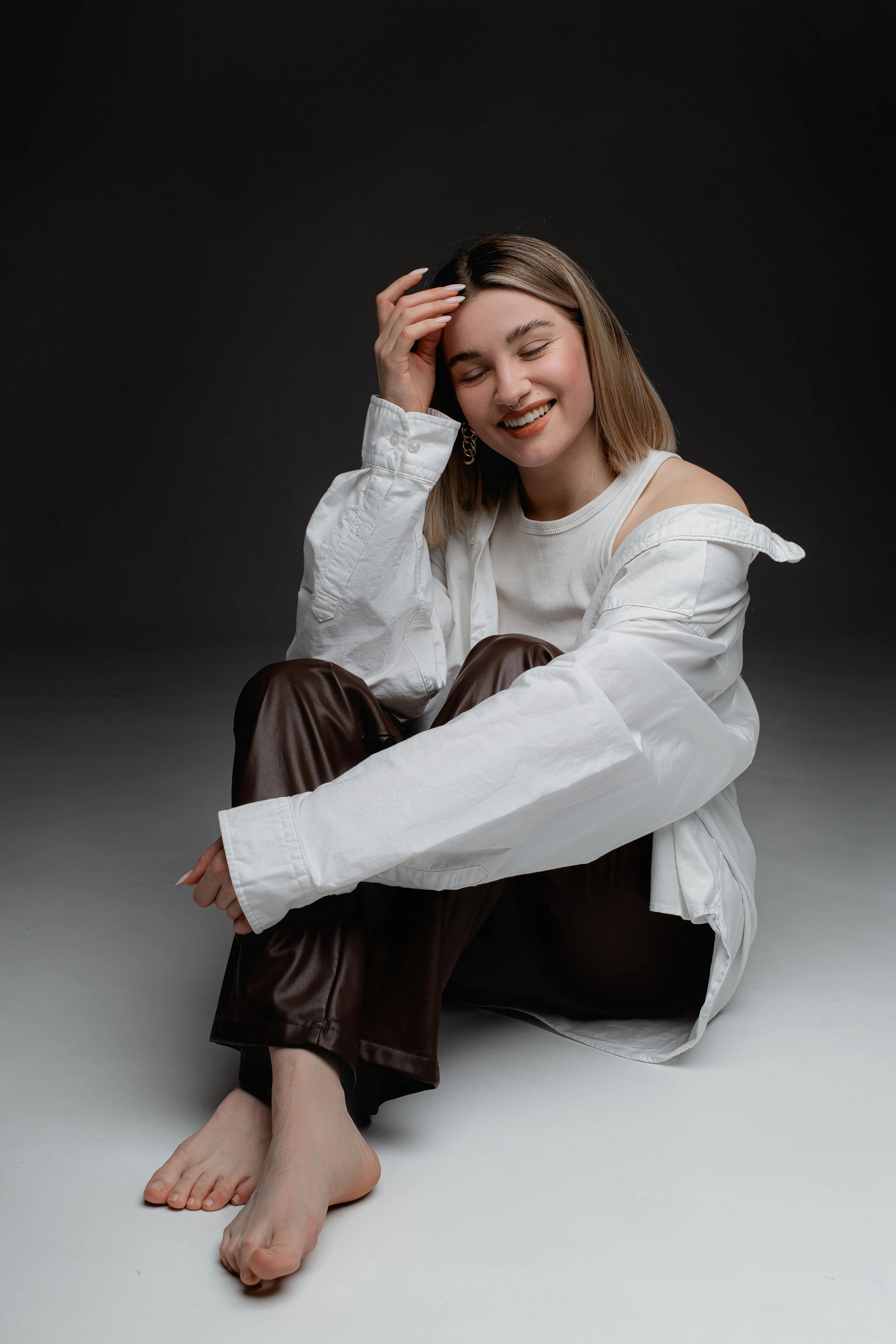 A young woman sitting barefoot on the floor, smiling with eyes closed, wearing a white top, a white oversized shirt, and brown pants, with her hand touching her forehead against a dark background.