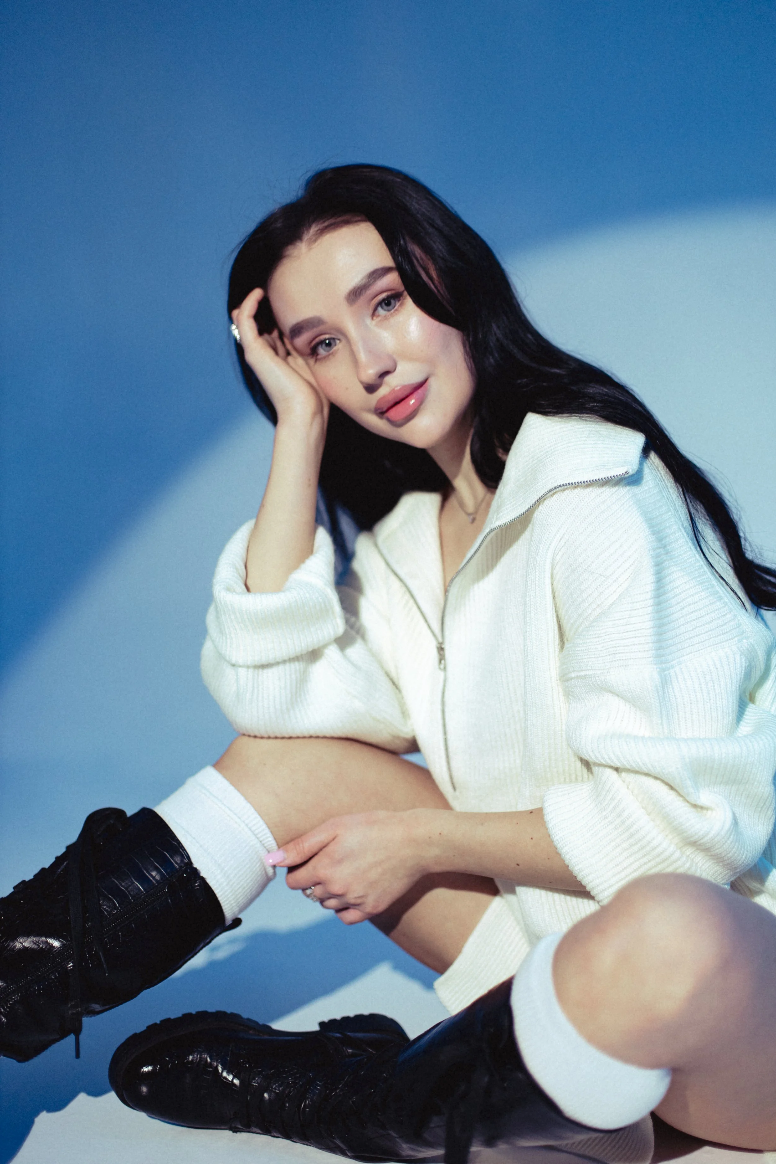 A young woman with long black hair and fair skin, dressed in a cream-colored zip-up sweater, is sitting with one knee up. She has a gentle smile and is resting her head on her left hand. She is wearing black lace-up boots, white socks, and a subtle n
