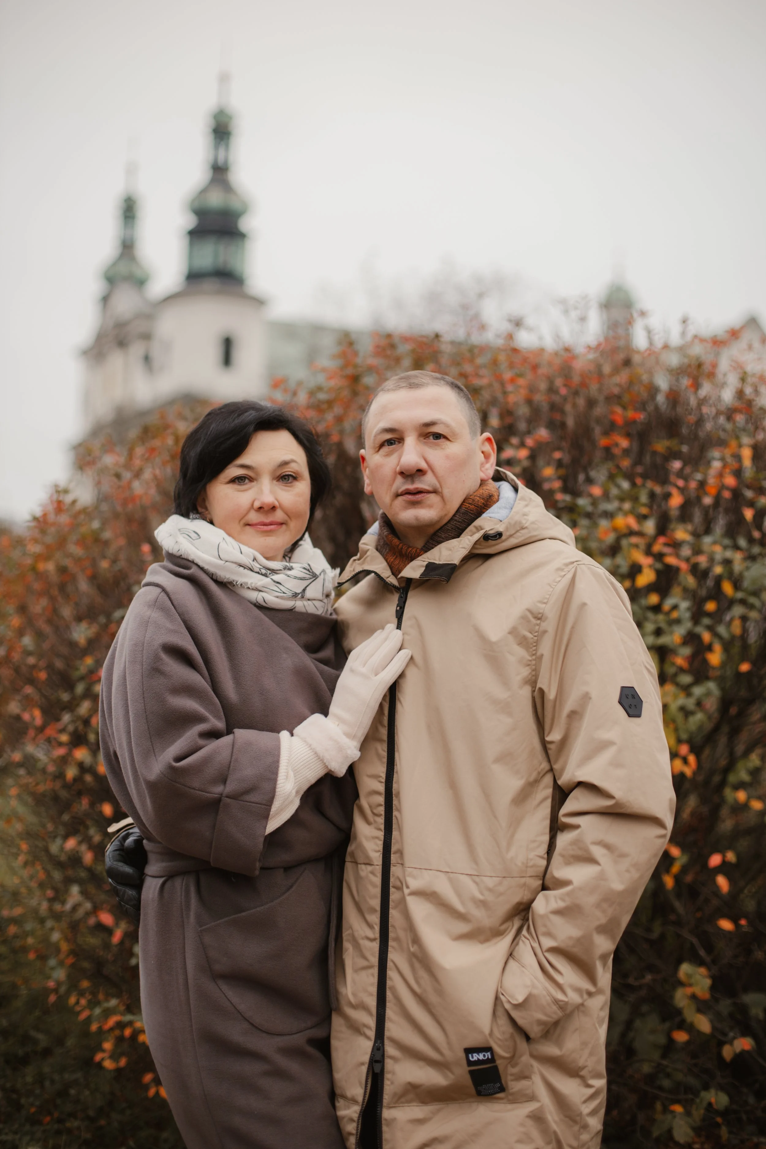 A middle-aged woman and man stand close together outdoors with a church steeple in the background, autumn foliage surrounds them.